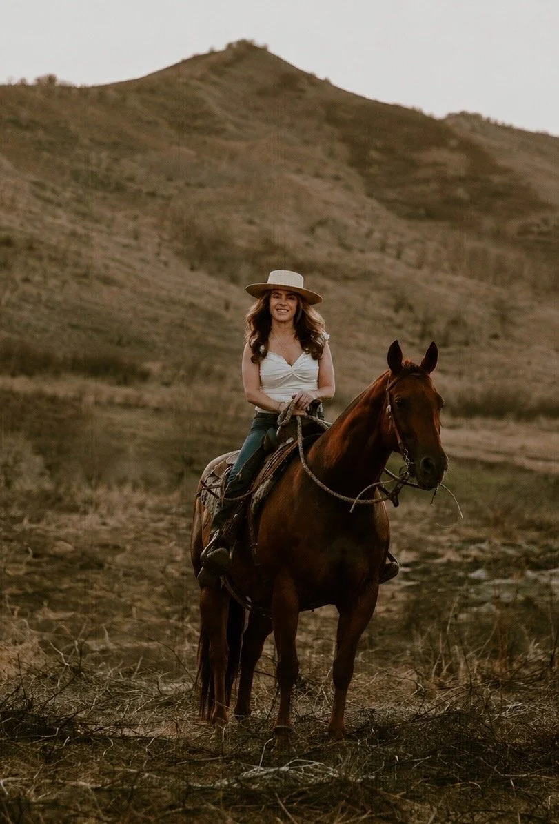 A woman's health coach wearing a white top and a wide-brimmed hat riding a brown horse in a rural, hilly landscape.