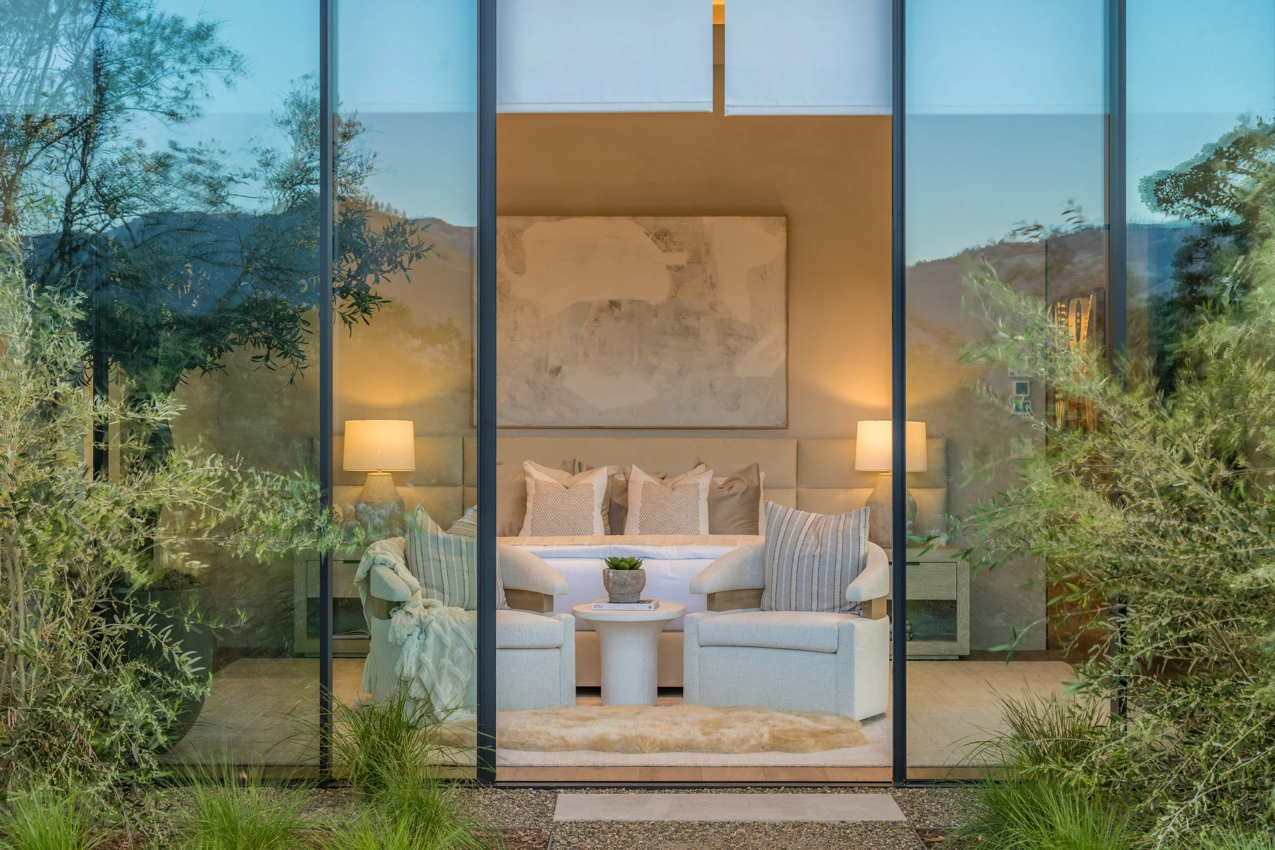 Living room with beige sofa, armchairs, and side tables, viewed through glass walls, with outdoor greenery