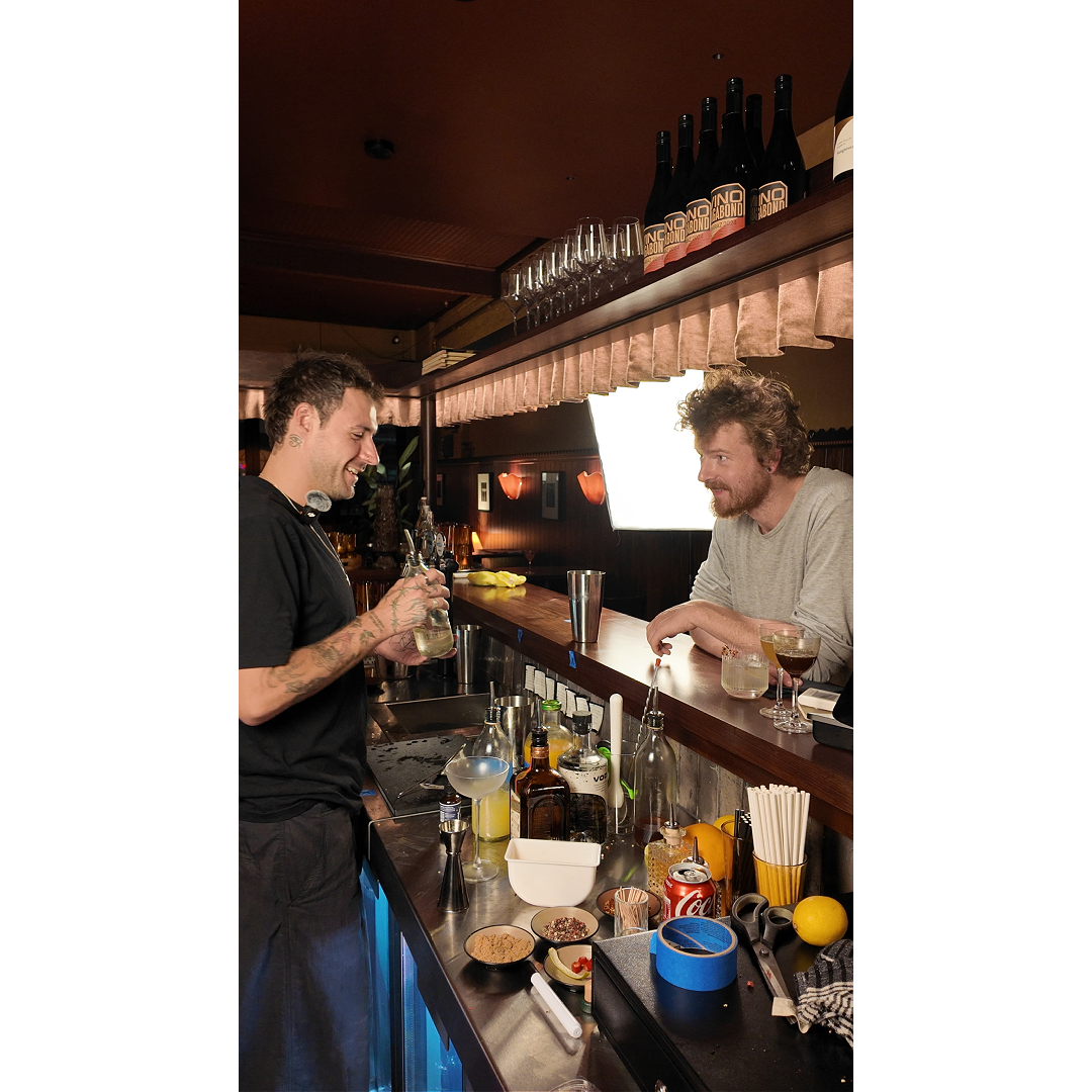 Two men at a bar, one behind the counter preparing a drink, and one preparing to video it, both smiling at each other. In the background is a professional photography light