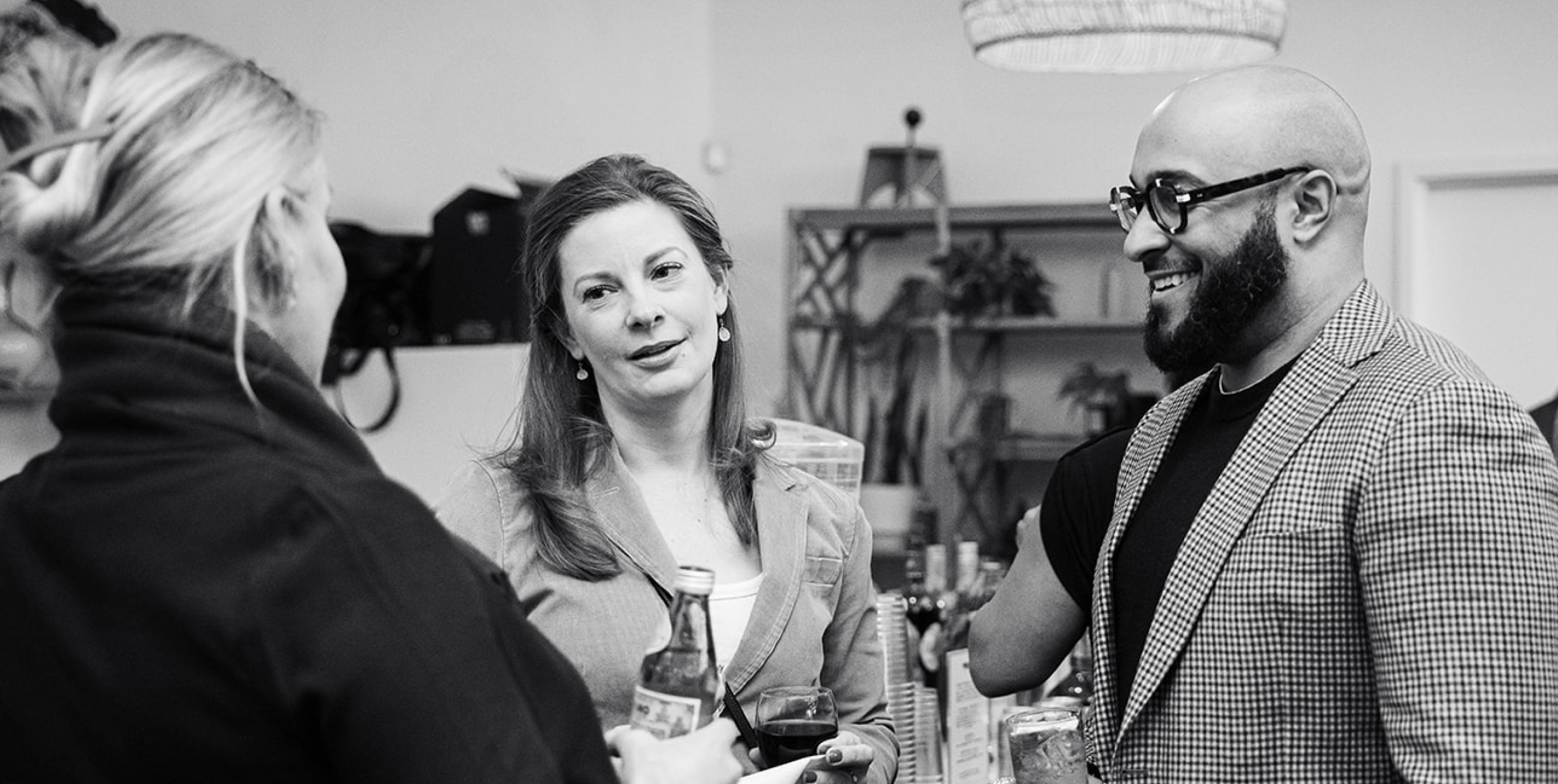 Four people are socializing at a bar or restaurant, engaging in conversation. One woman is speaking, and a man with glasses and a beard is smiling. The scene is in black and white.