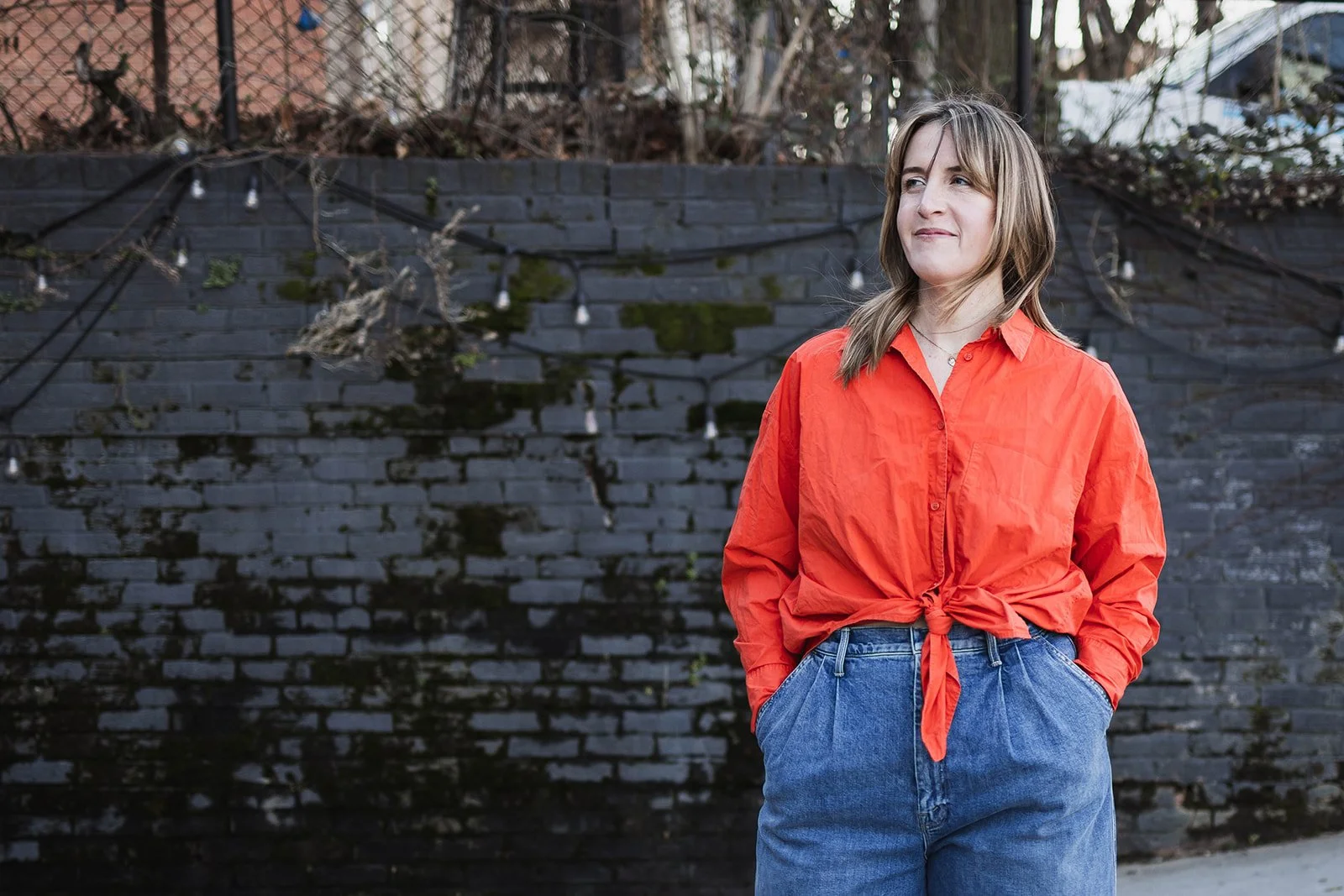 A woman with shoulder-length light brown hair wearing a red tied-up shirt and blue jeans standing outdoors against a dark brick wall with string lights.