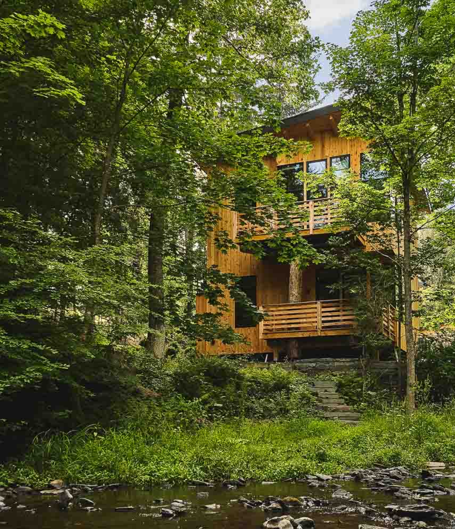 A wooden house with multiple balconies is surrounded by lush green trees in a forest setting, with a small stream in the foreground.