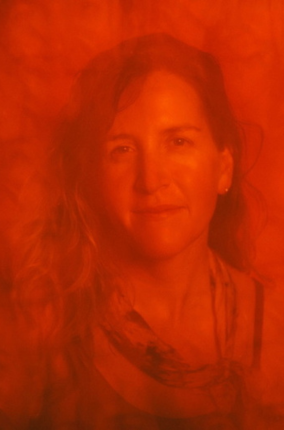 Portrait of a woman with shoulder-length hair, wearing a scarf, illuminated by warm, reddish lighting.