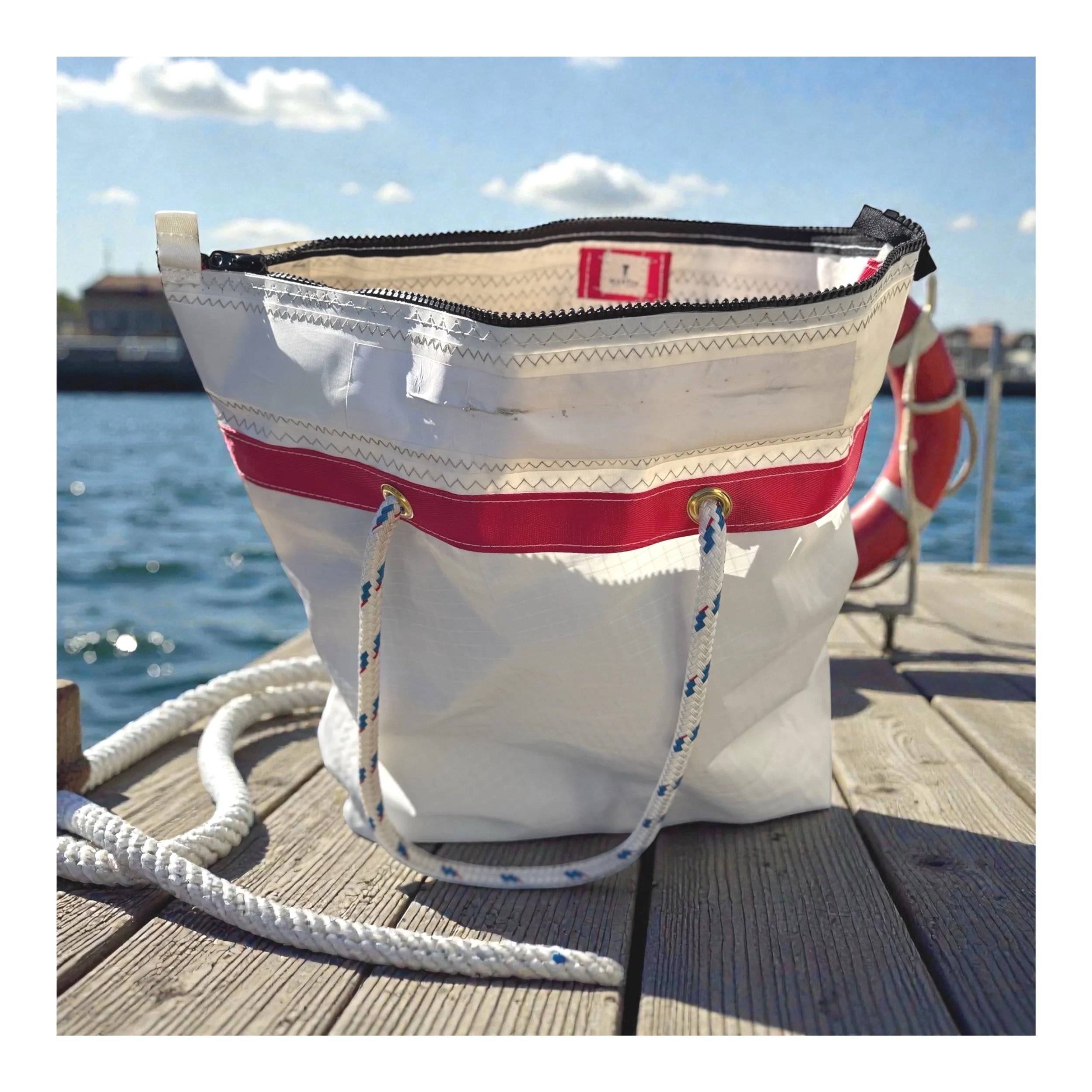 Close-up of a white and red fabric bag with a black zipper on a wooden dock near water under a partly cloudy sky, with ropes in the foreground.
