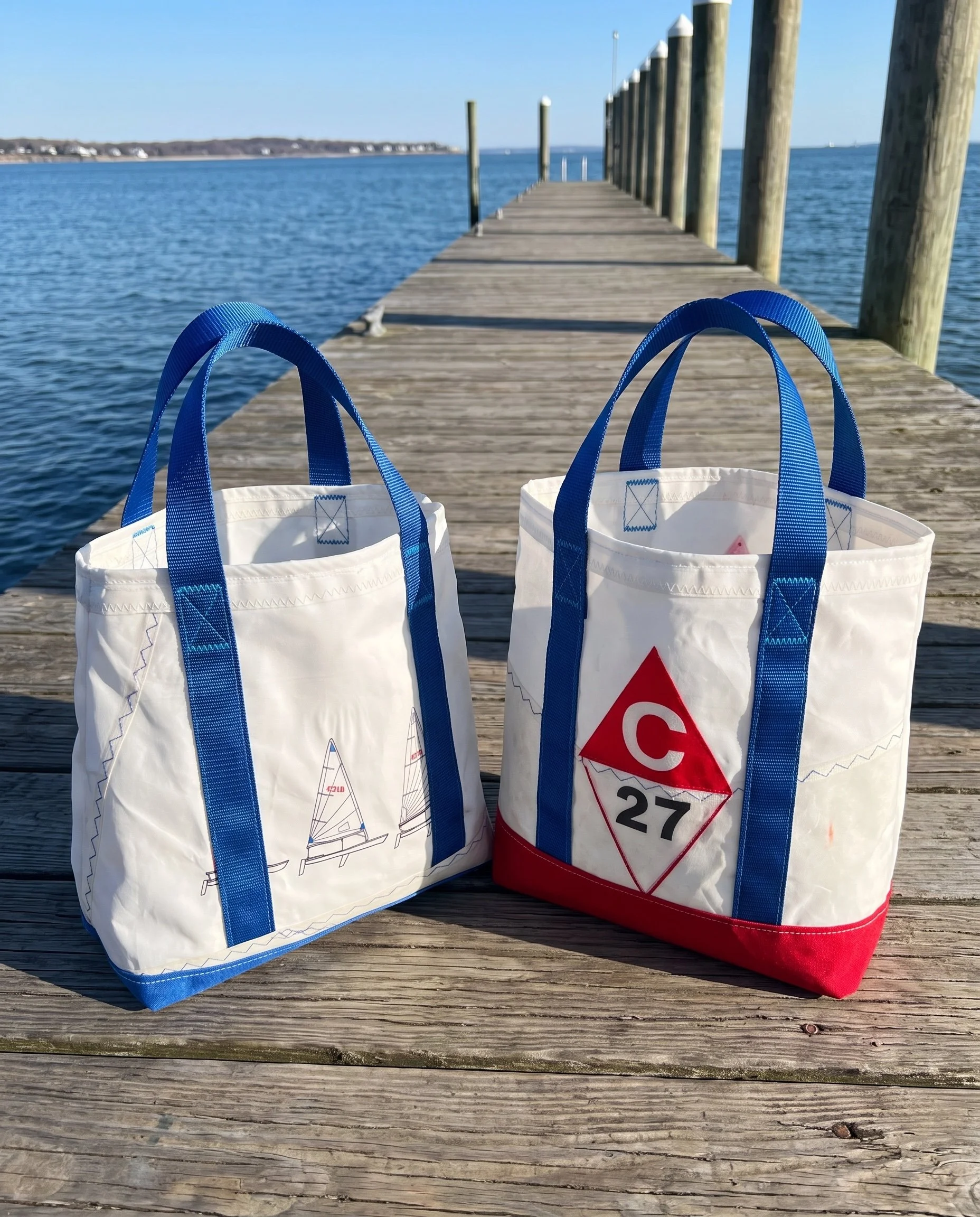 Two white tote bags with blue handles placed on a wooden dock overlooking a body of water, with a distant shoreline and a clear blue sky in the background.