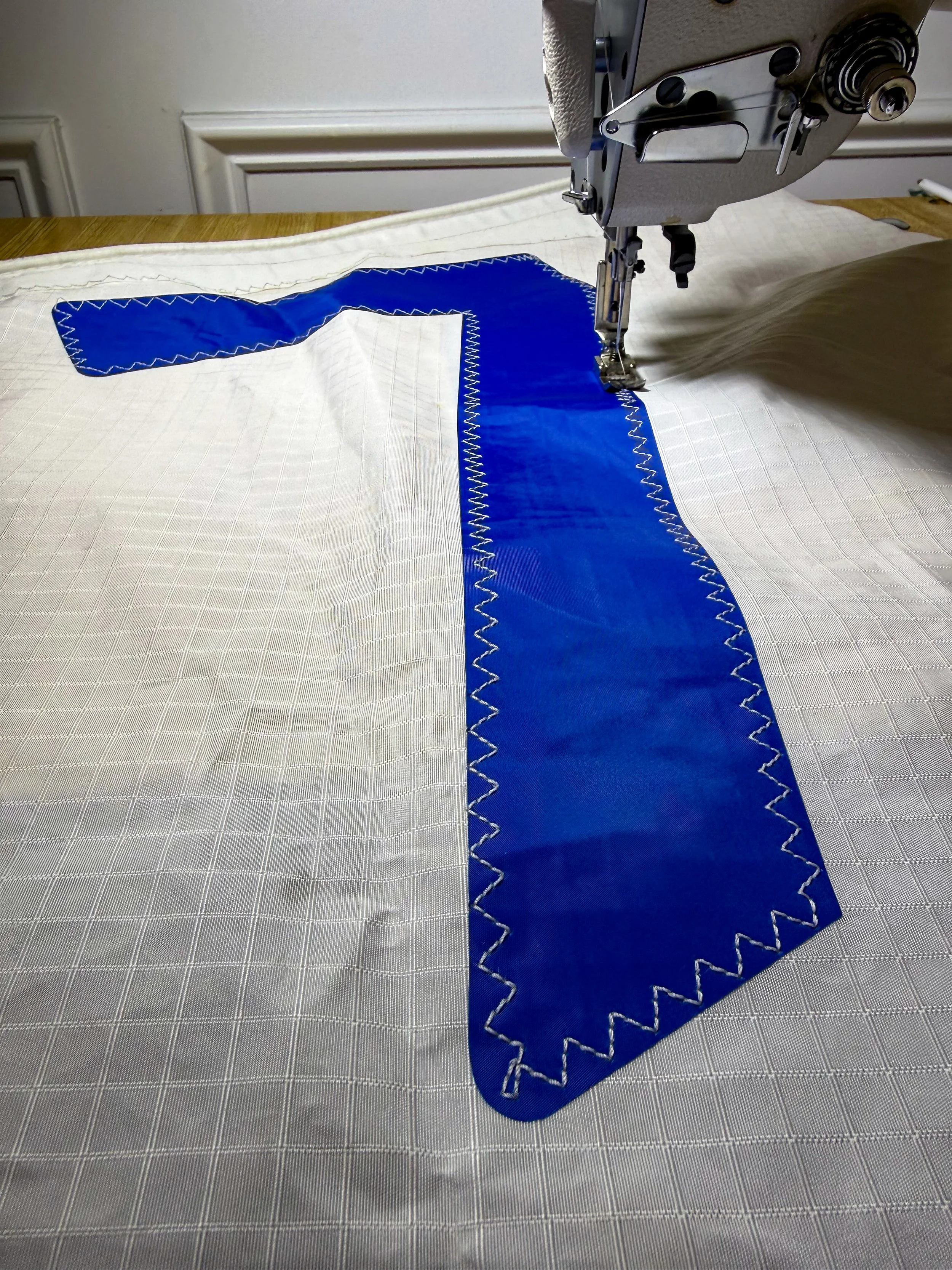 Close-up of a sewing machine stitching a blue fabric onto a white fabric with a grid pattern. The blue fabric is shaped like a letter 'T'.
