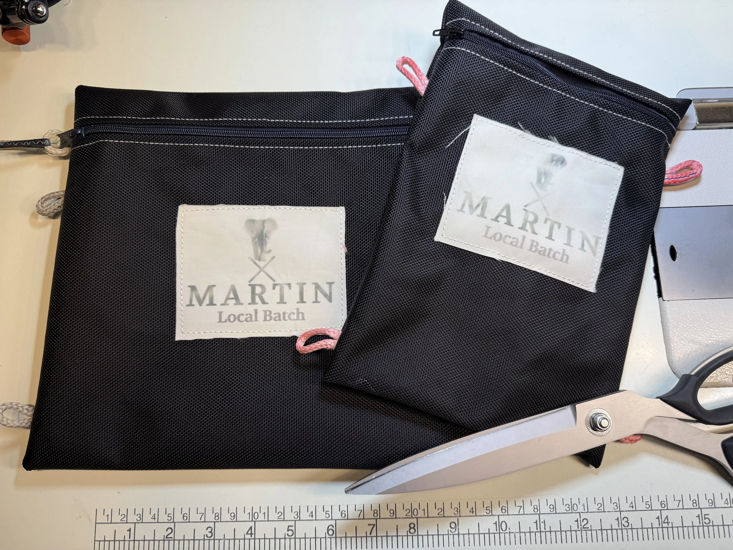 Two black fabric pouches with white stitching and pink loop pulls, placed on a metal surface next to a ruler for measurement.