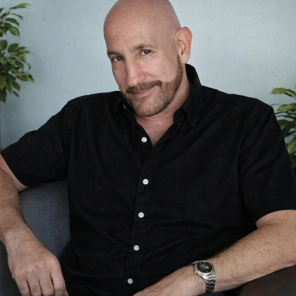 A man with a shaved head and beard wearing a black button-up shirt sitting on a gray couch with plants in the background.