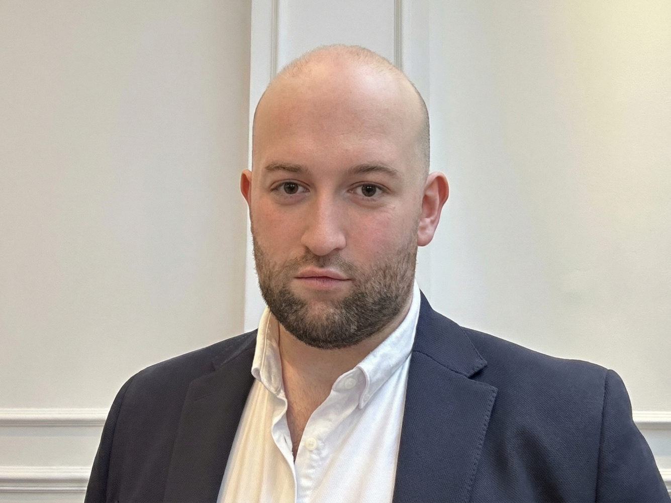 A young man with a shaved head and beard wearing a white collared shirt and a dark blazer, standing indoors in front of a plain light-colored wall.