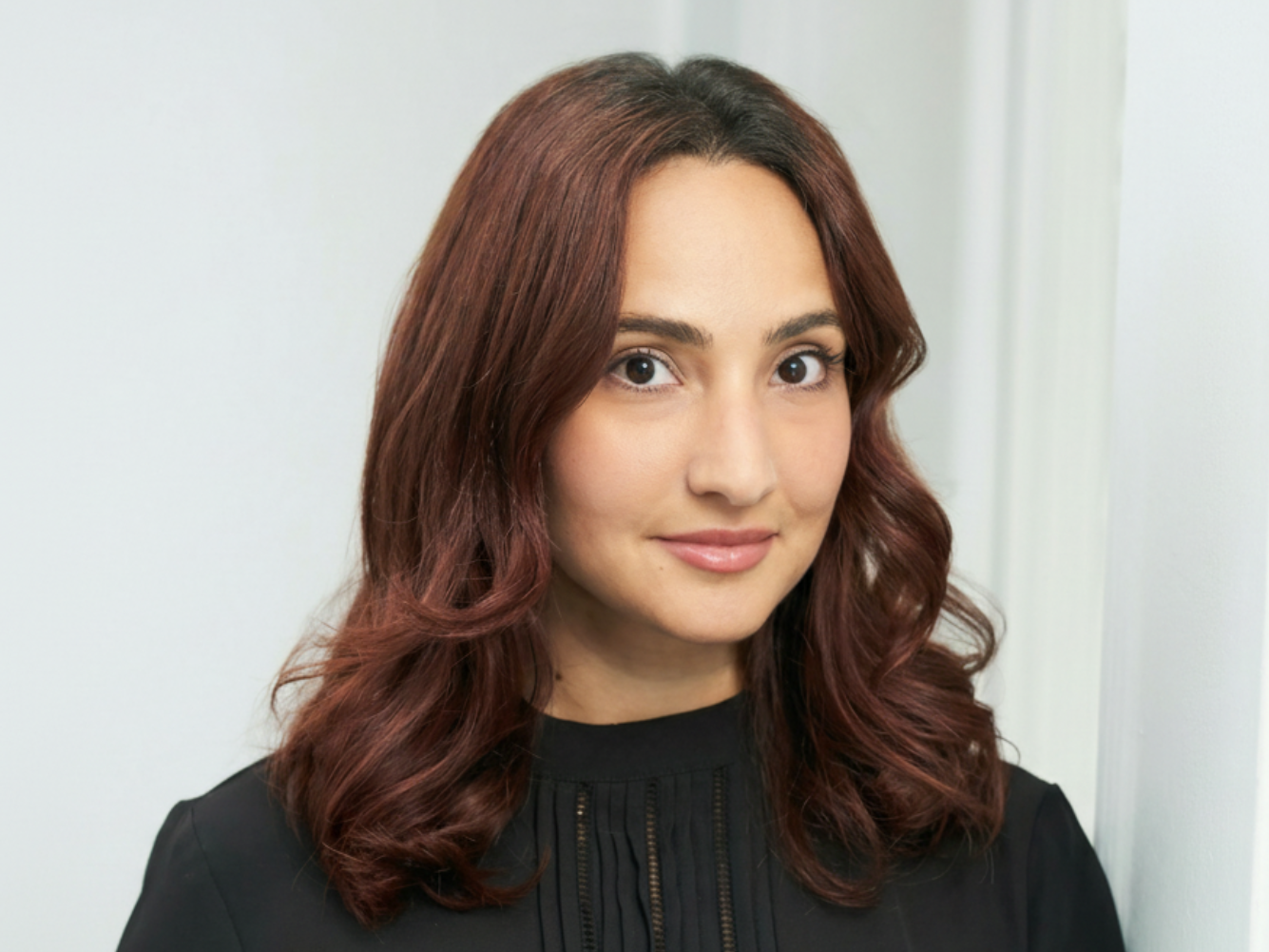 A woman with medium skin, shoulder-length auburn hair styled in loose waves, wearing a black top, standing in front of a plain white wall.