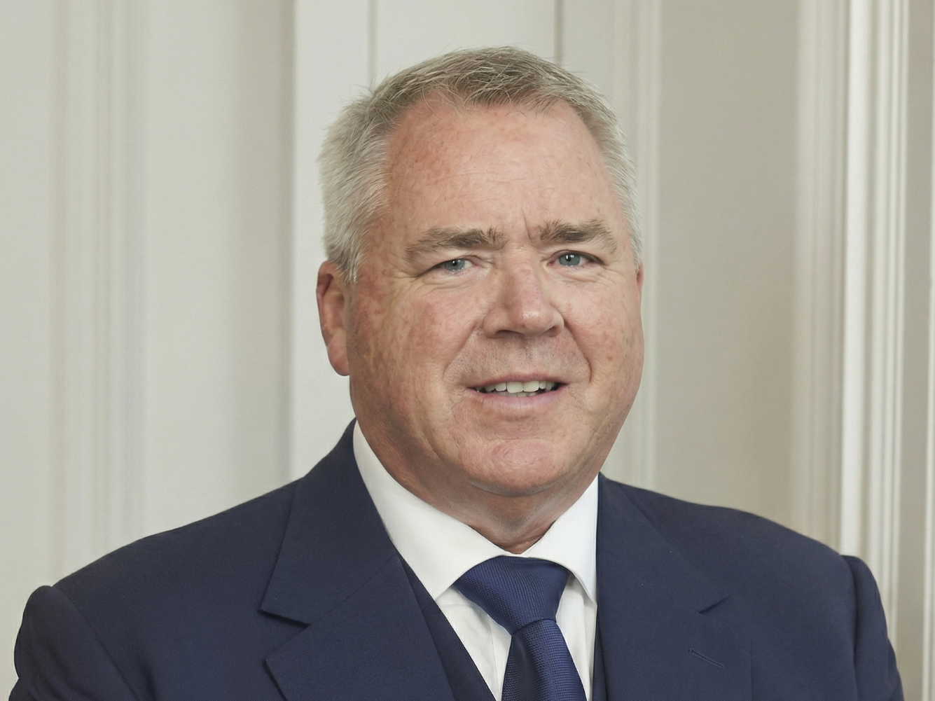 A middle-aged man with gray hair, blue eyes, and wearing a dark suit, white shirt, and navy tie, standing in front of a light-colored wooden wall.