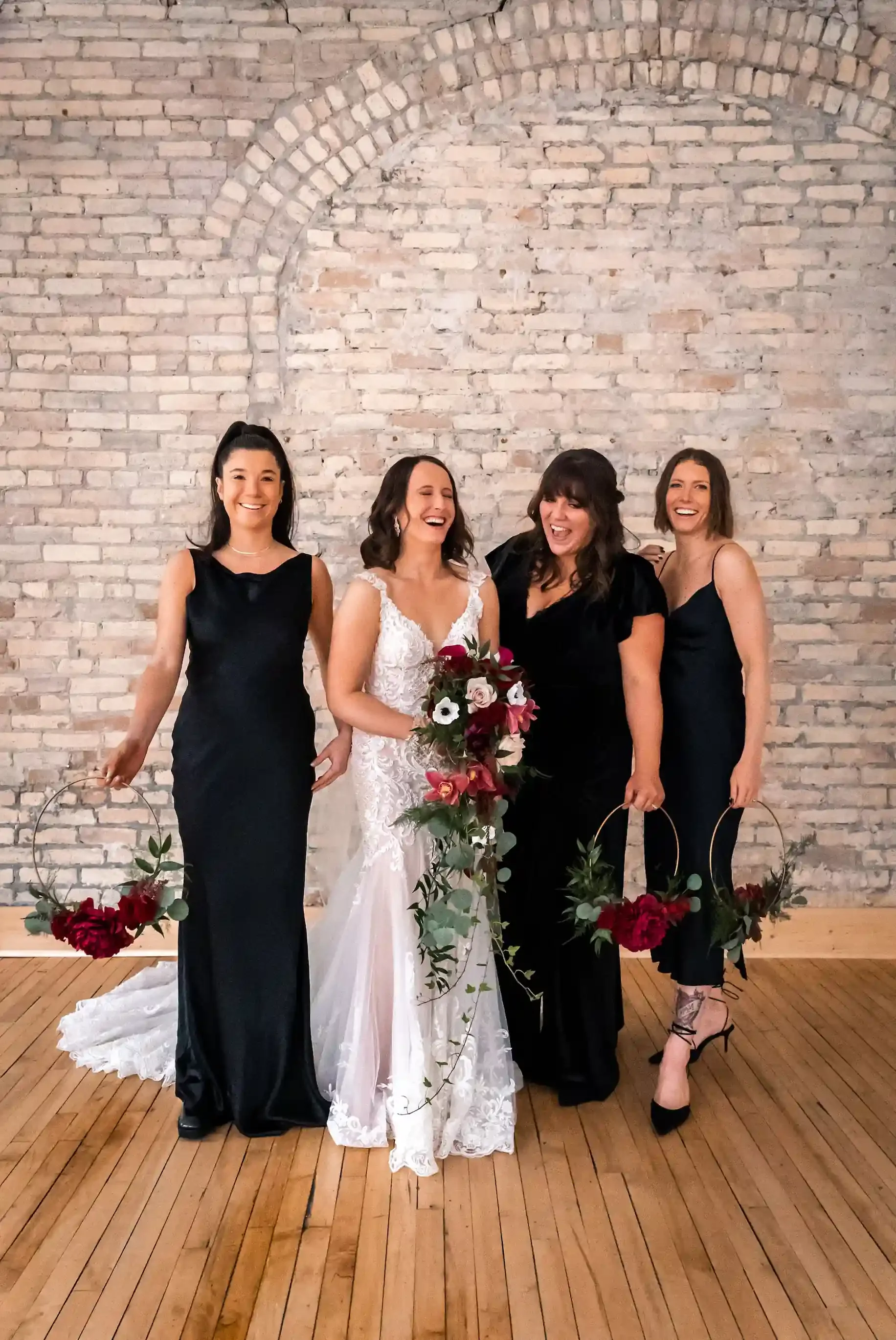 Bride and bridesmaids laughing together in front of the exposed brick arch wall at The Harlow Exchange wedding venue in Winnipeg's Exchange District