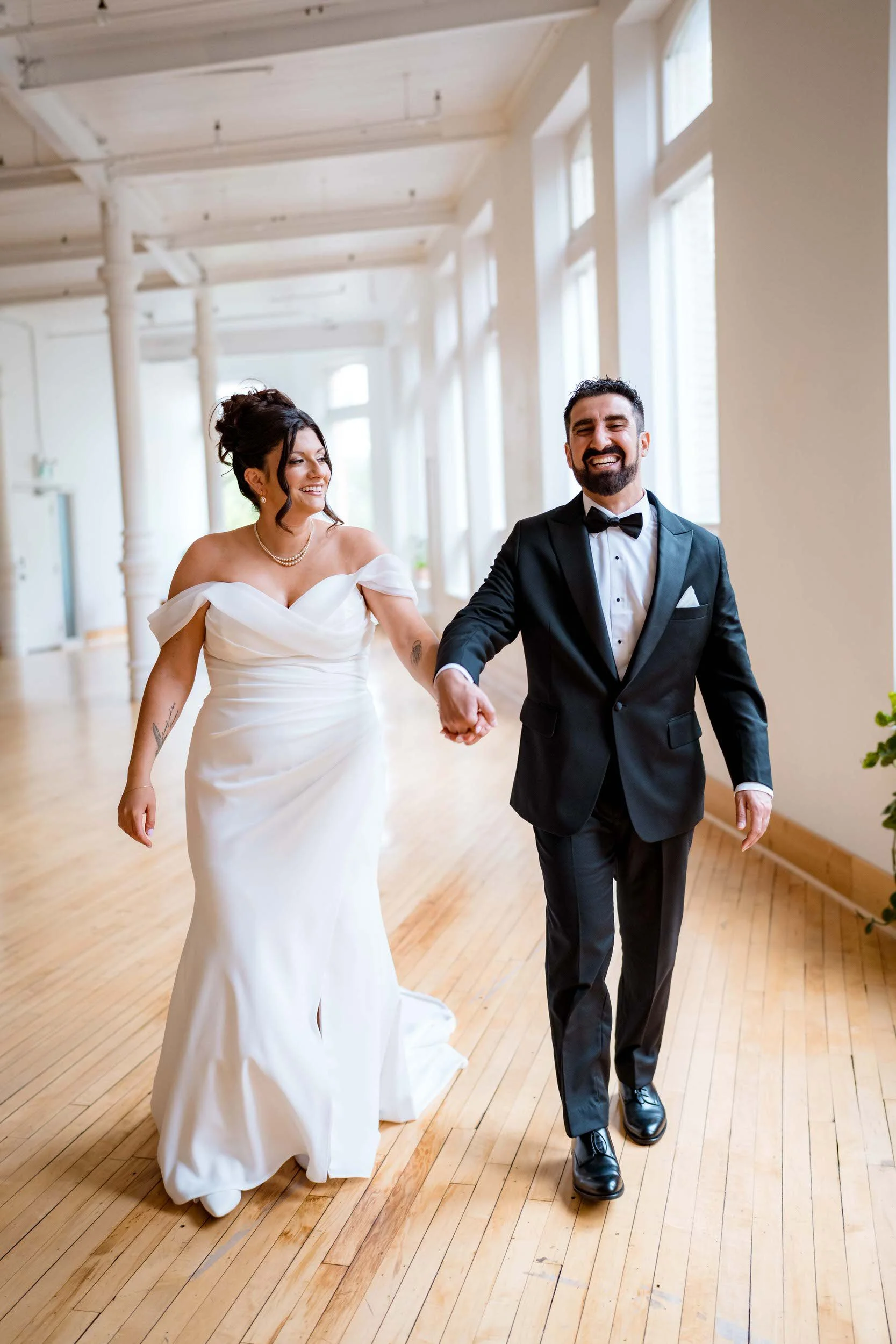 Couple walking through The Harlow Exchange event space on their wedding day — hardwood floors, white columns, natural window light