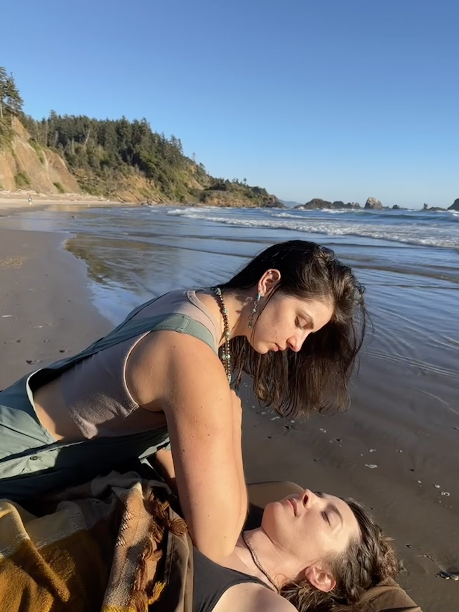 A woman and a girl are lying on a sandy beach, with the woman comforting the girl. The ocean waves are gently lapping at the shore, and a cliff with trees can be seen in the background under a clear blue sky.