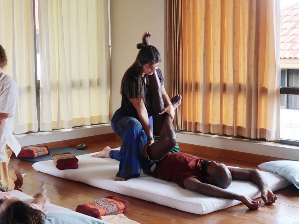 A woman practices Thai massage on a man lying on a mattress in a sunlit room with curtains.