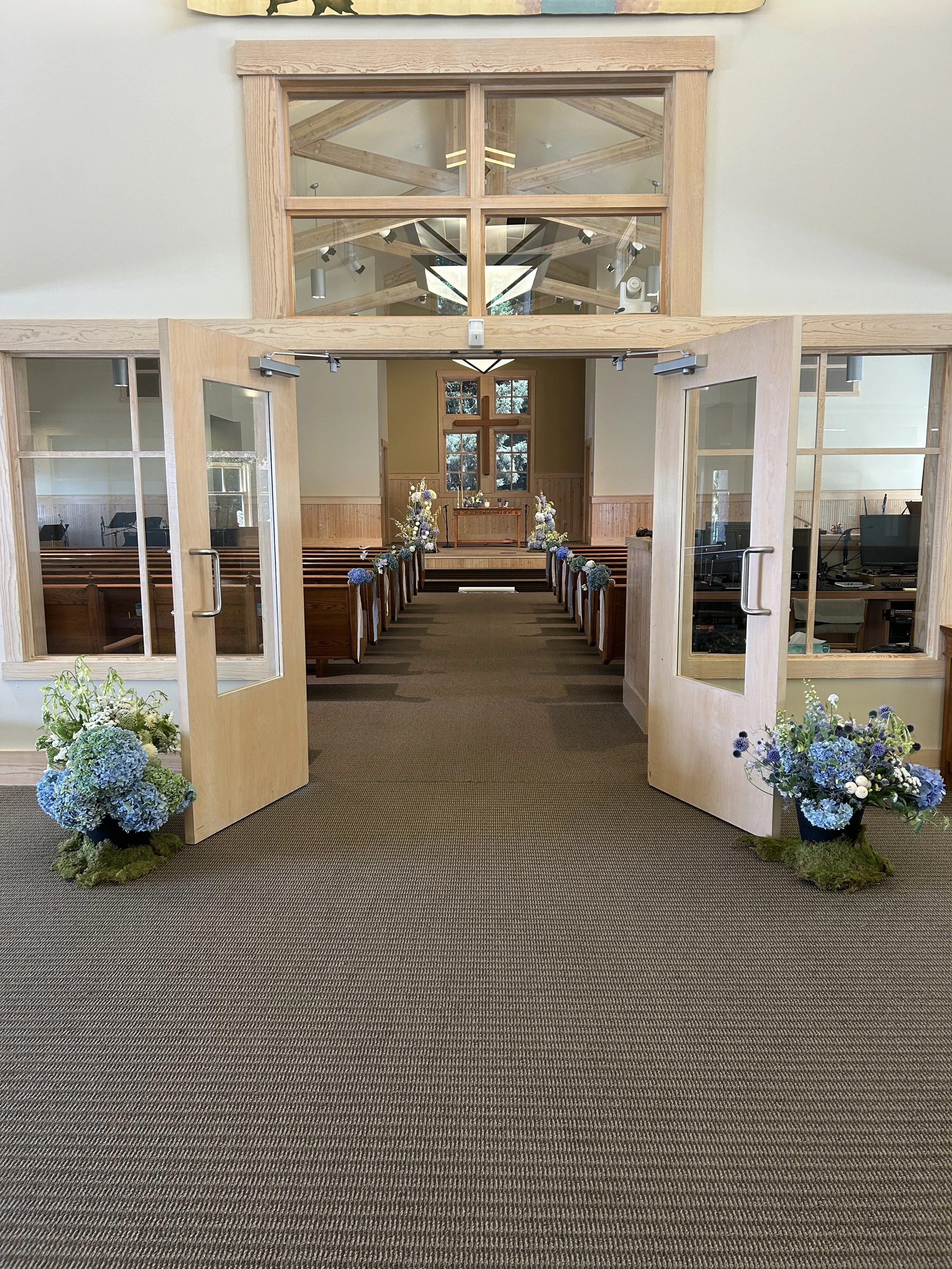 Open double doors with flower arrangements on each side leading into an empty chapel decorated with flowers, with pews and altar at the front.