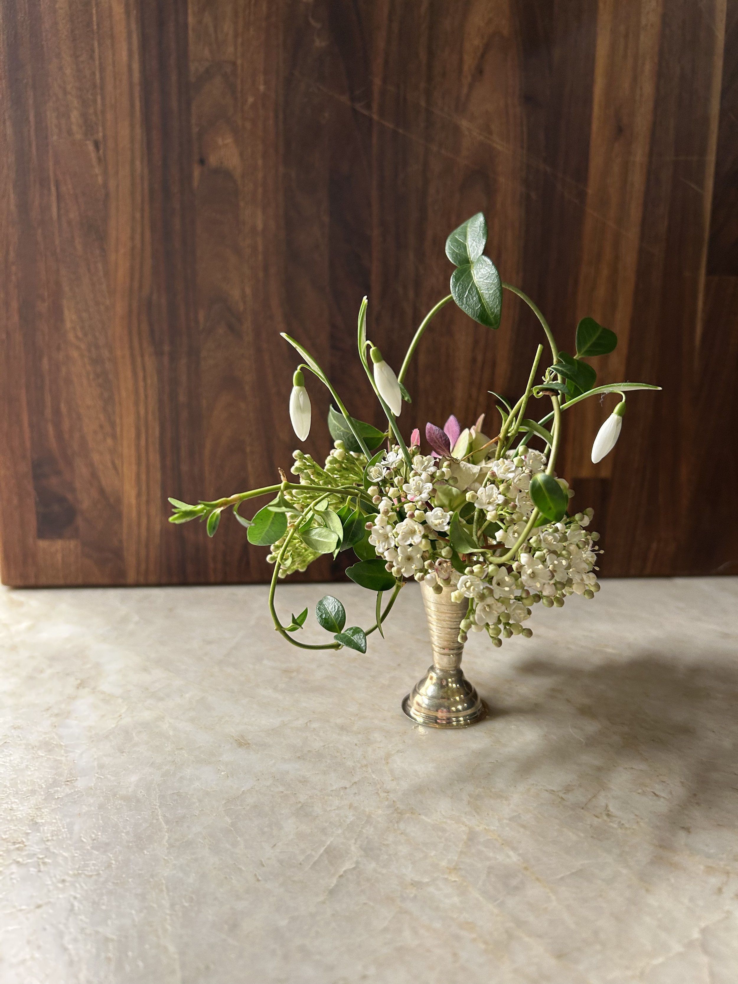 Small metal vase filled with white and pink flowers, green leaves, and sprigs of greenery on a beige surface with a wooden background.