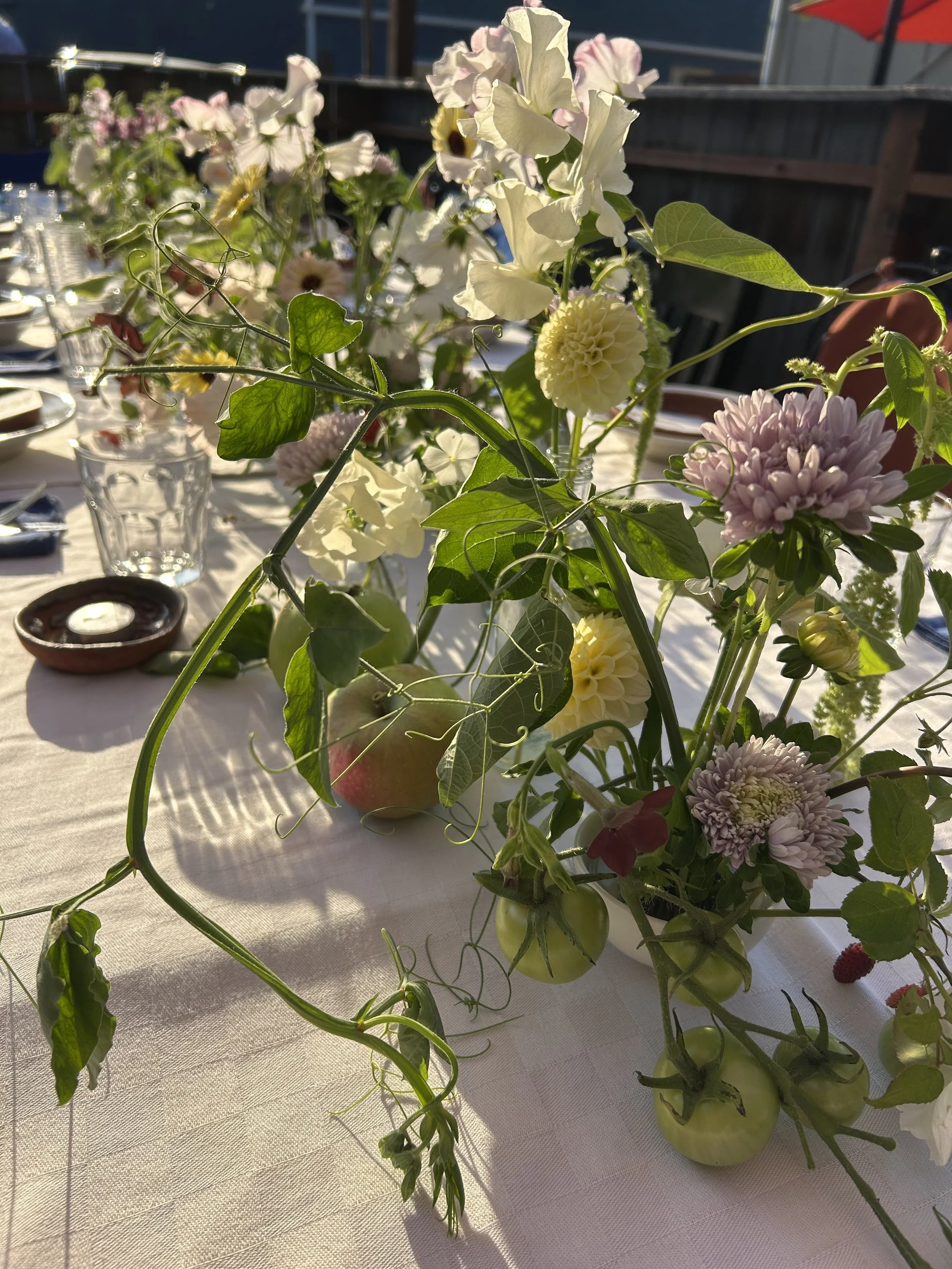 A floral arrangement with various colorful flowers, tomatoes, and greenery on a table, with sunlight casting shadows and a blurred outdoor background.