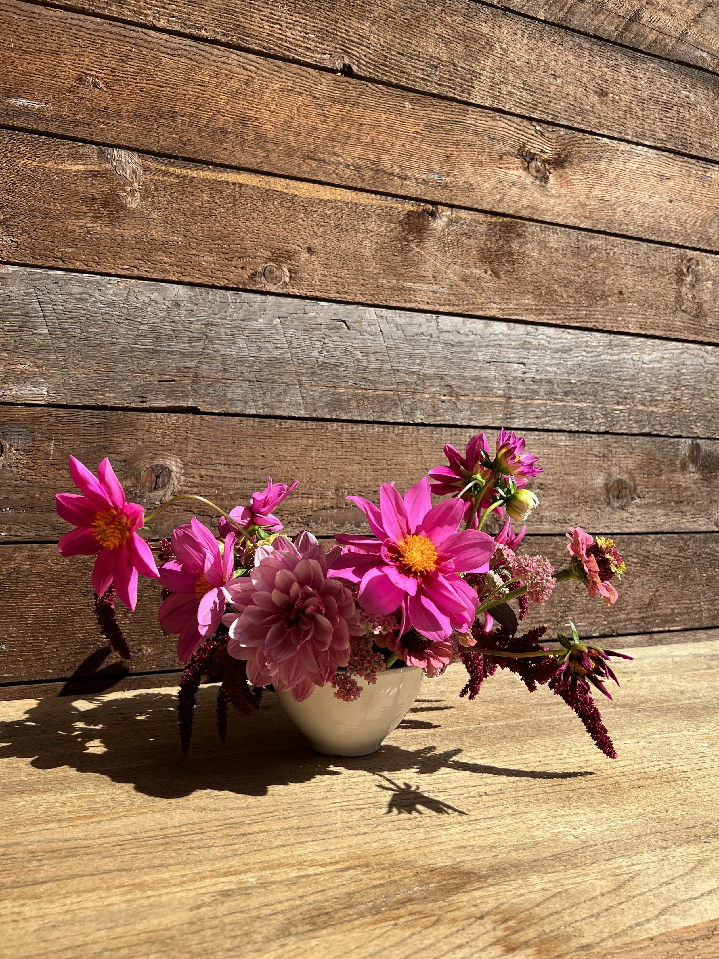 A small white ceramic vase holding an arrangement of pink flowers, including dahlias and other blooms, on a wooden surface with a wooden wall background.