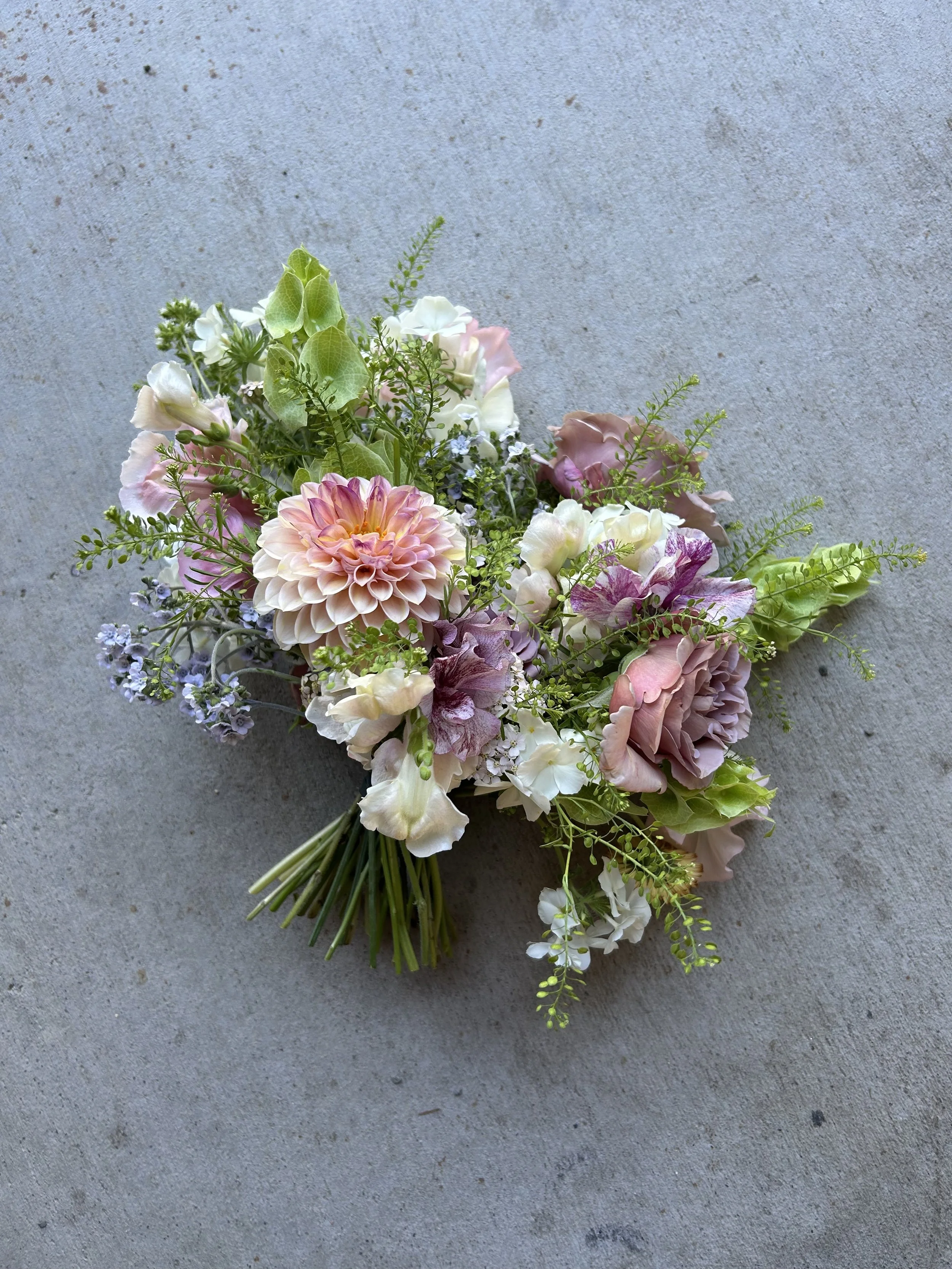 A bridal bouquet of mixed flowers on a gray surface, including pink, white, and purple blooms with green foliage.