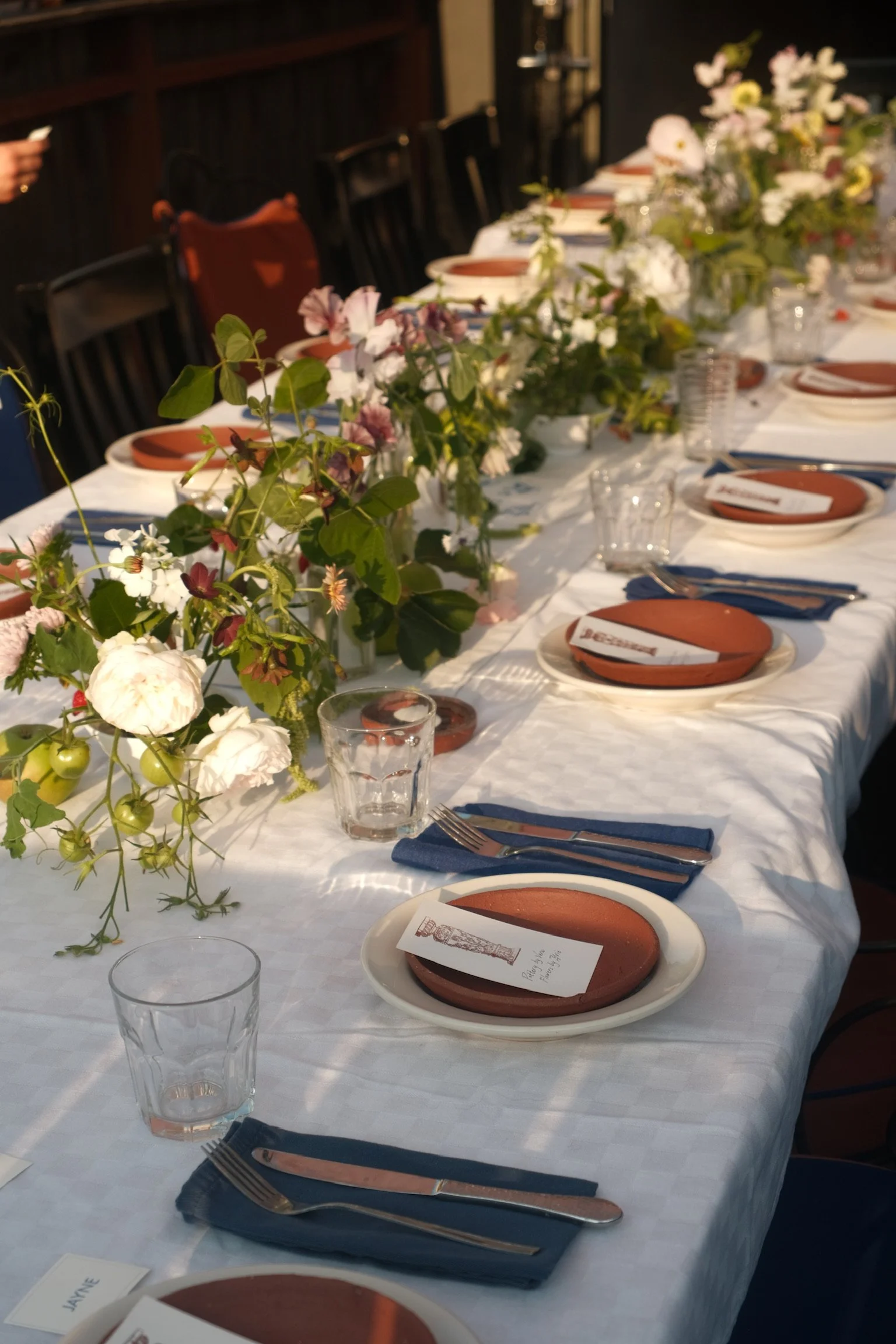 A long dining table set for a meal with plates, forks, knives, drinking glasses, and floral centerpieces, with sunlight casting shadows on the table.