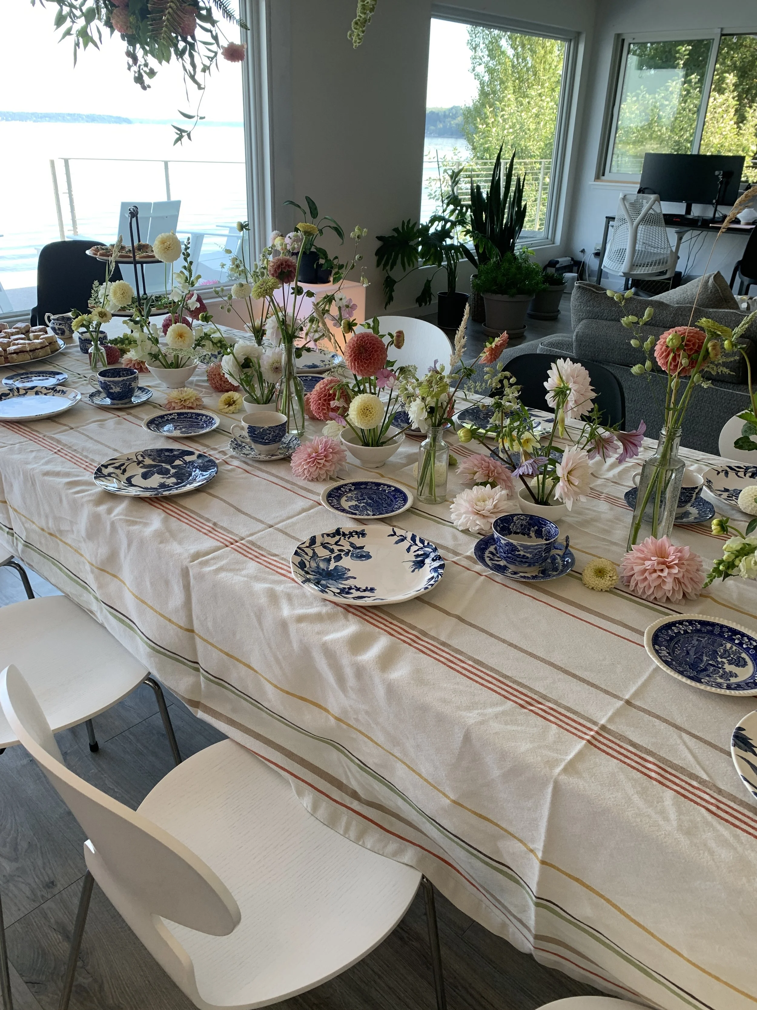 Table set with blue and white plates, cups, and teapot, decorated with pink and white flowers in vases, near large windows overlooking water and greenery.