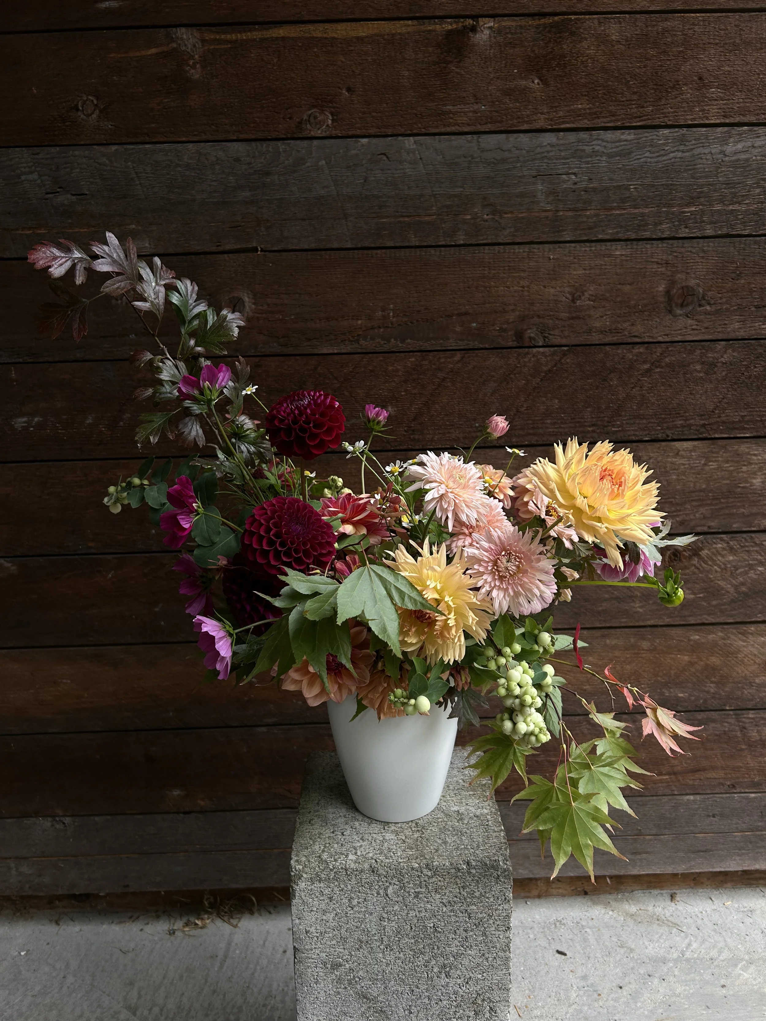A bouquet of colorful flowers in a white vase placed on a concrete block against a wooden wall.