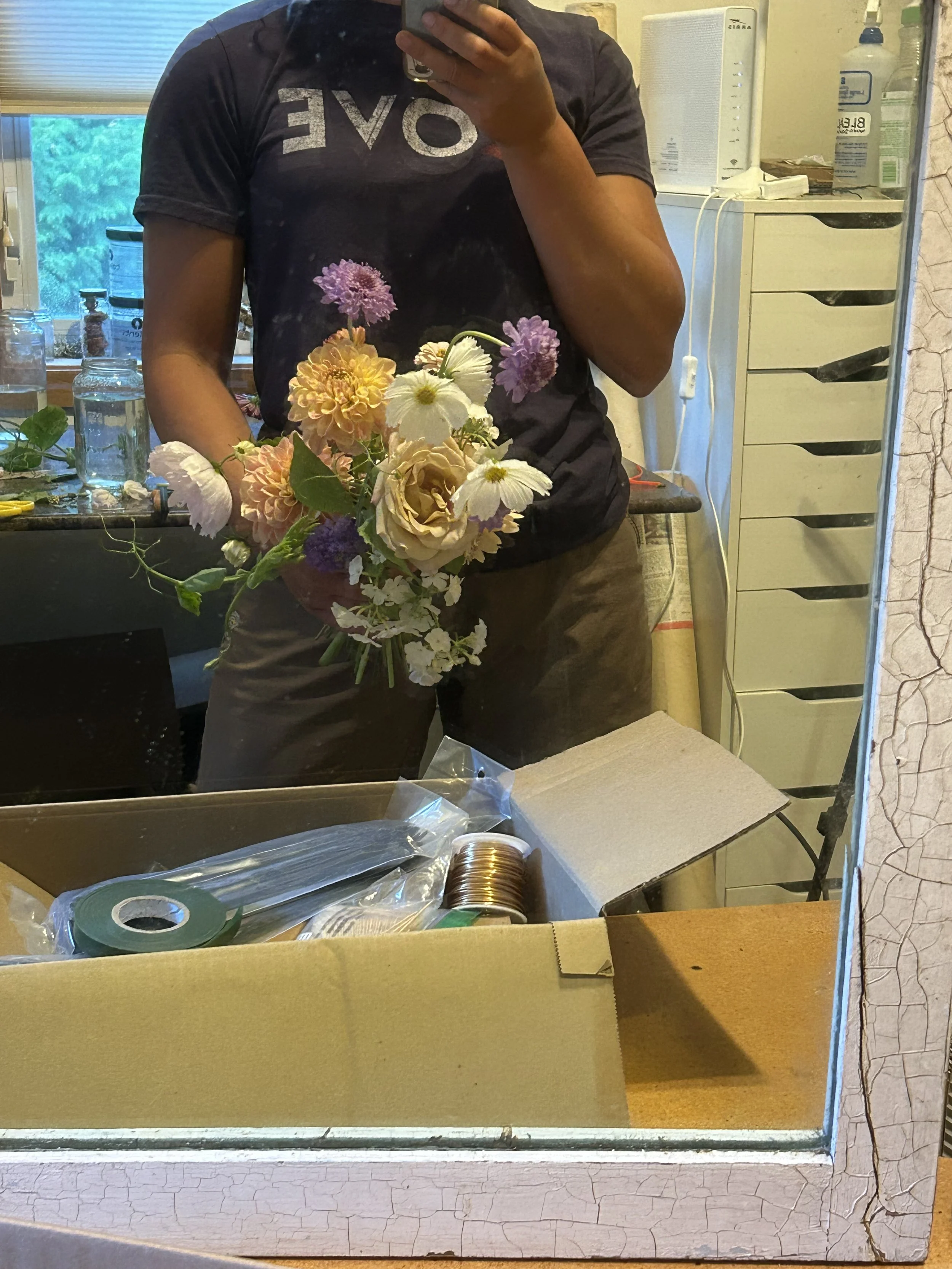 First person taking a mirror selfie holding a bouquet of colorful flowers, with a cluttered workspace and drawer cabinet in the background.