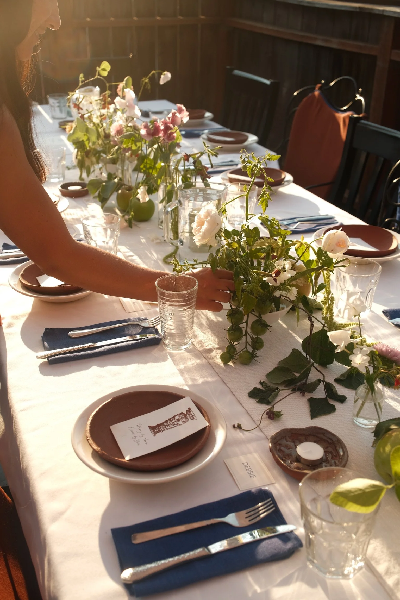 A long dining table set for a meal with plates, silverware, glasses, and centerpieces of flowers. One person is placing a floral arrangement on the table, and the setting includes a menu or place card. The scene is lit by natural light, creating a wa