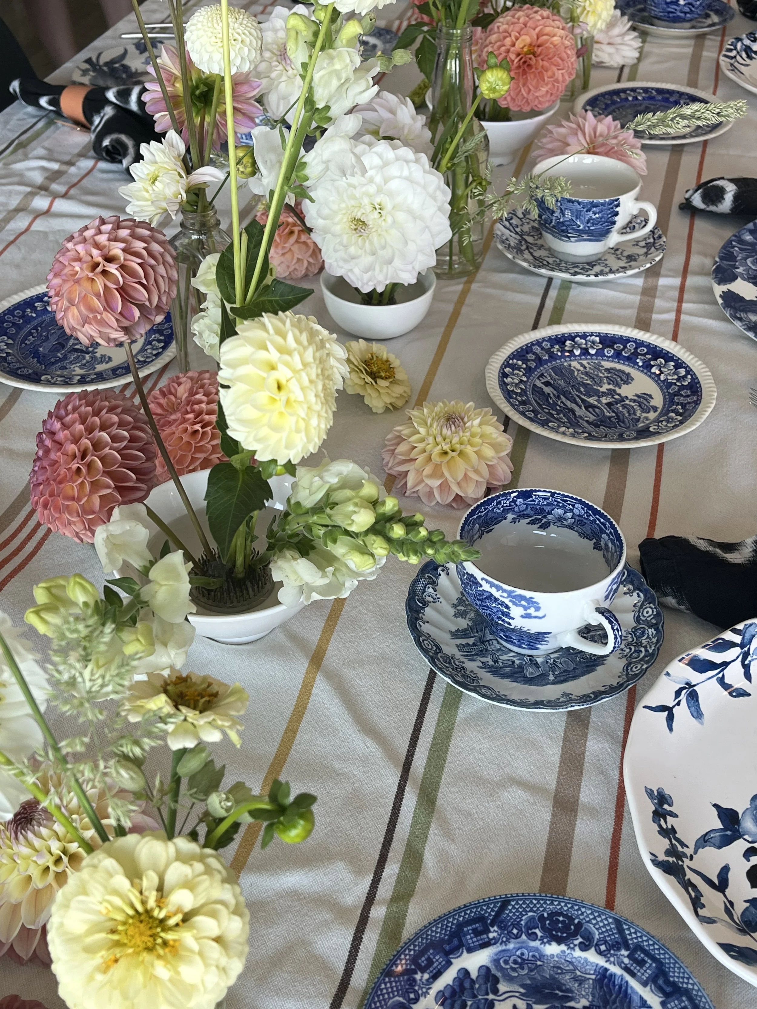 A dining table decorated with multiple vases of colorful flowers and set with blue and white patterned porcelain dishes, teacups, and plates, with some black and white napkins.