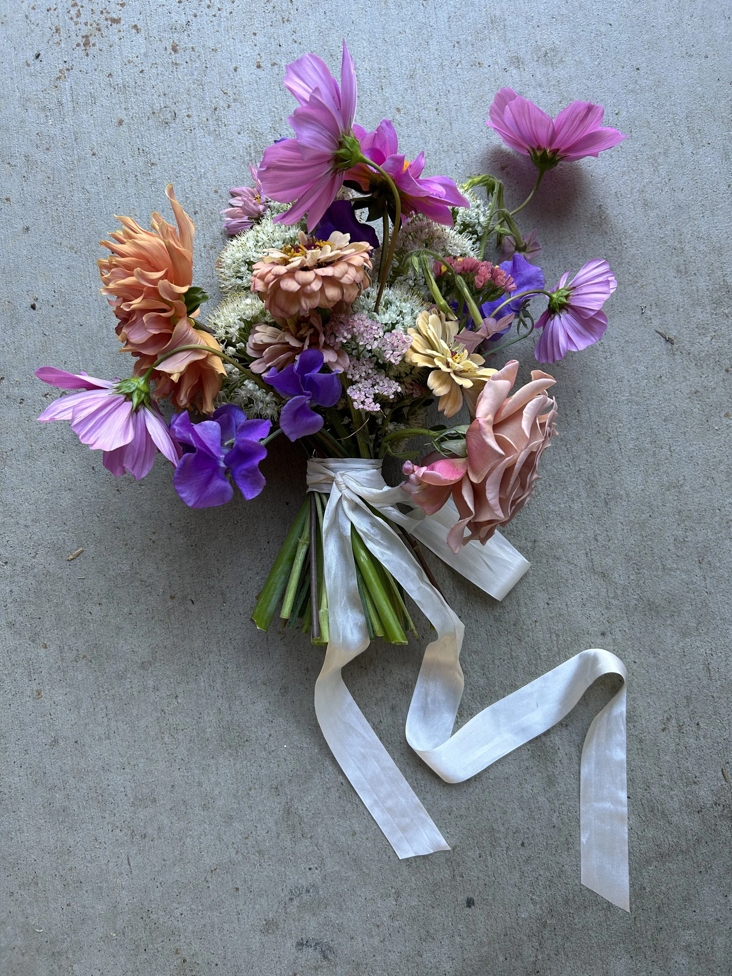A colorful bouquet of various flowers tied with a white ribbon, placed on a concrete surface.