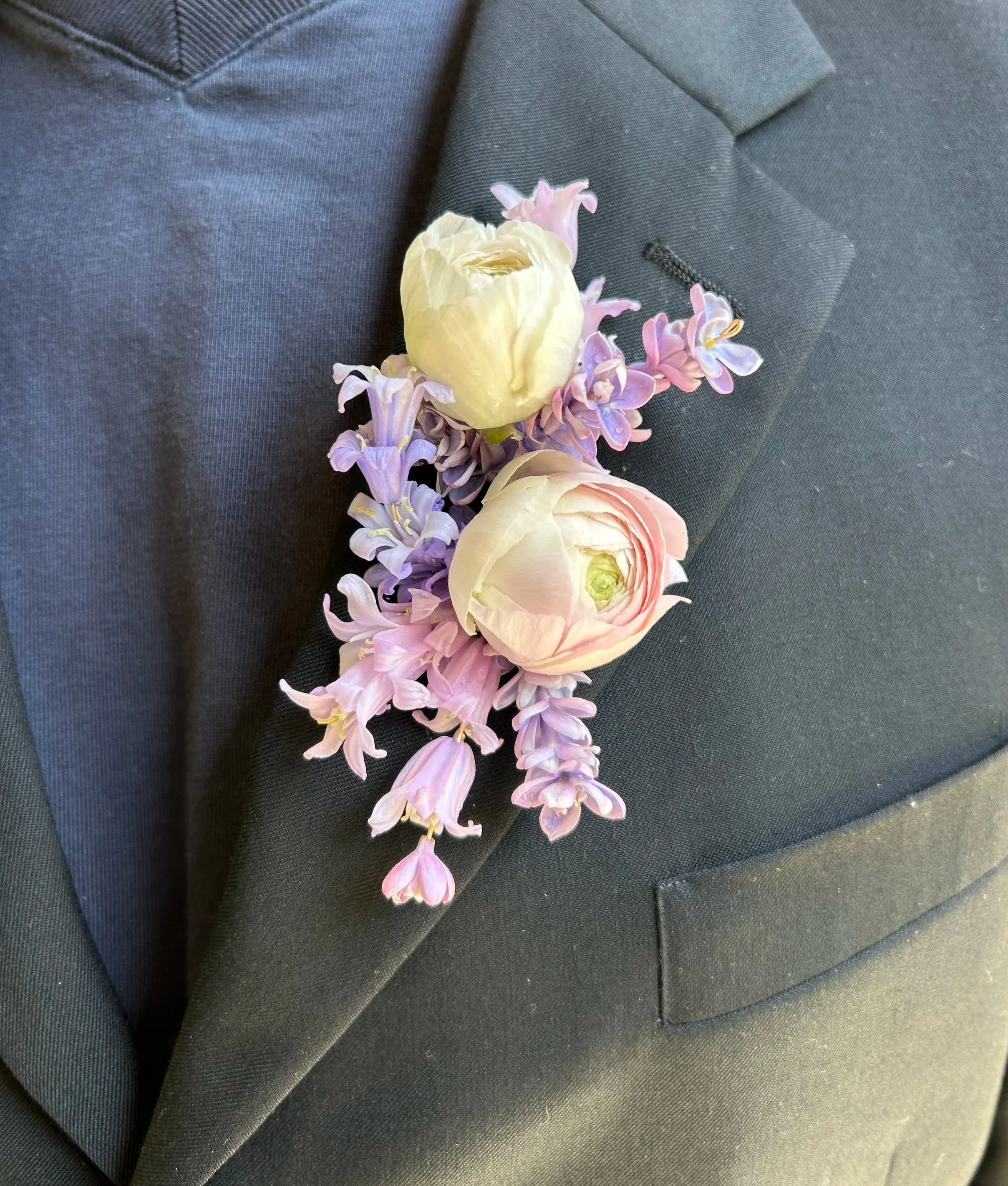 A boutonnière floral arrangement with white and pink ranunculus and purple hyacinth, pinned to a dark gray suit jacket.