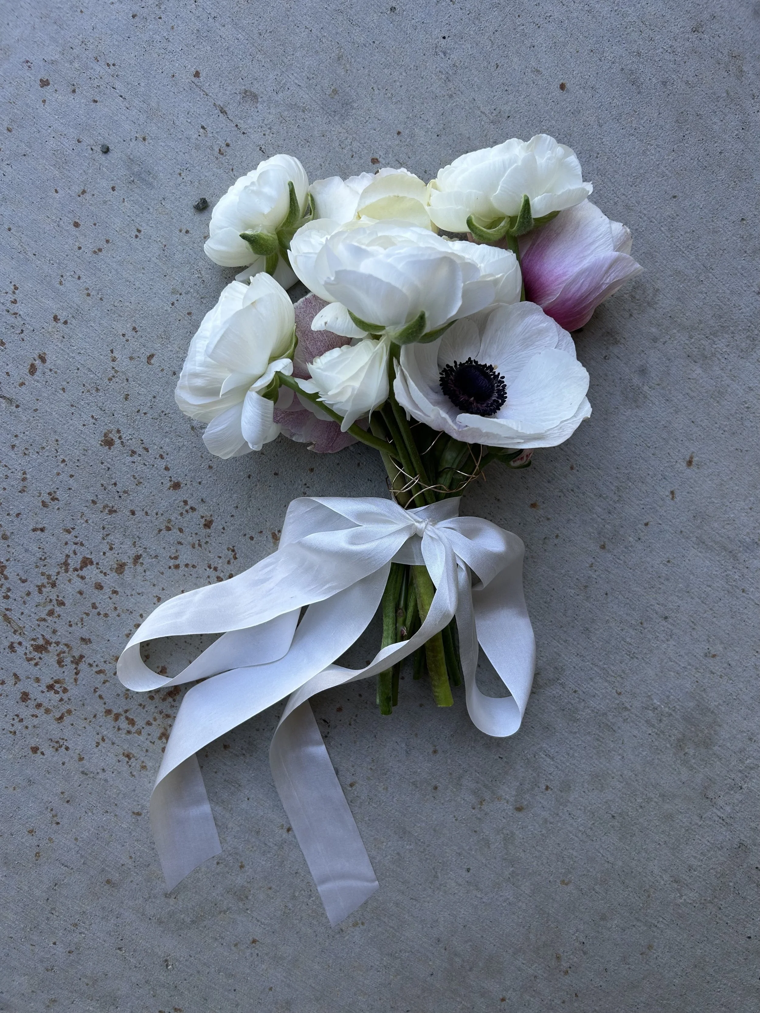 A small bouquet of white anemone and ranunculus flowers with a pink bud, tied with a white ribbon, resting on a gray concrete surface.