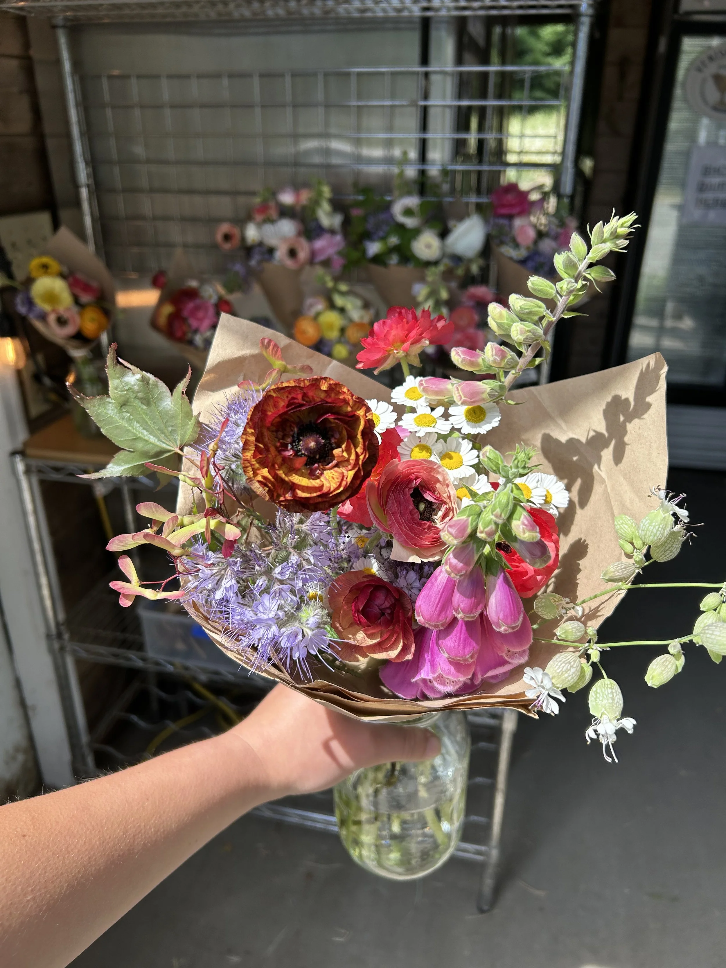 Hand holding a glass jar with a colorful bouquet of mixed flowers including pink, red, purple, and white blooms in front of a flower shop with additional bouquets on display.