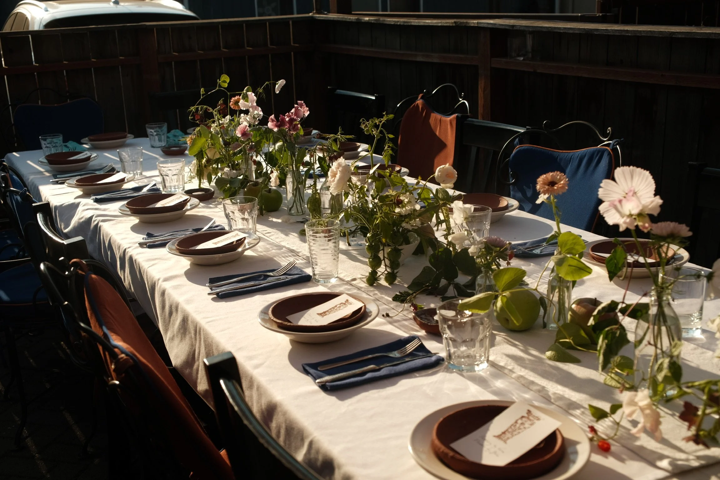 A long outdoor dining table set with plates, glasses, and napkins, decorated with a flower arrangement in the center, on a patio with wooden fencing.