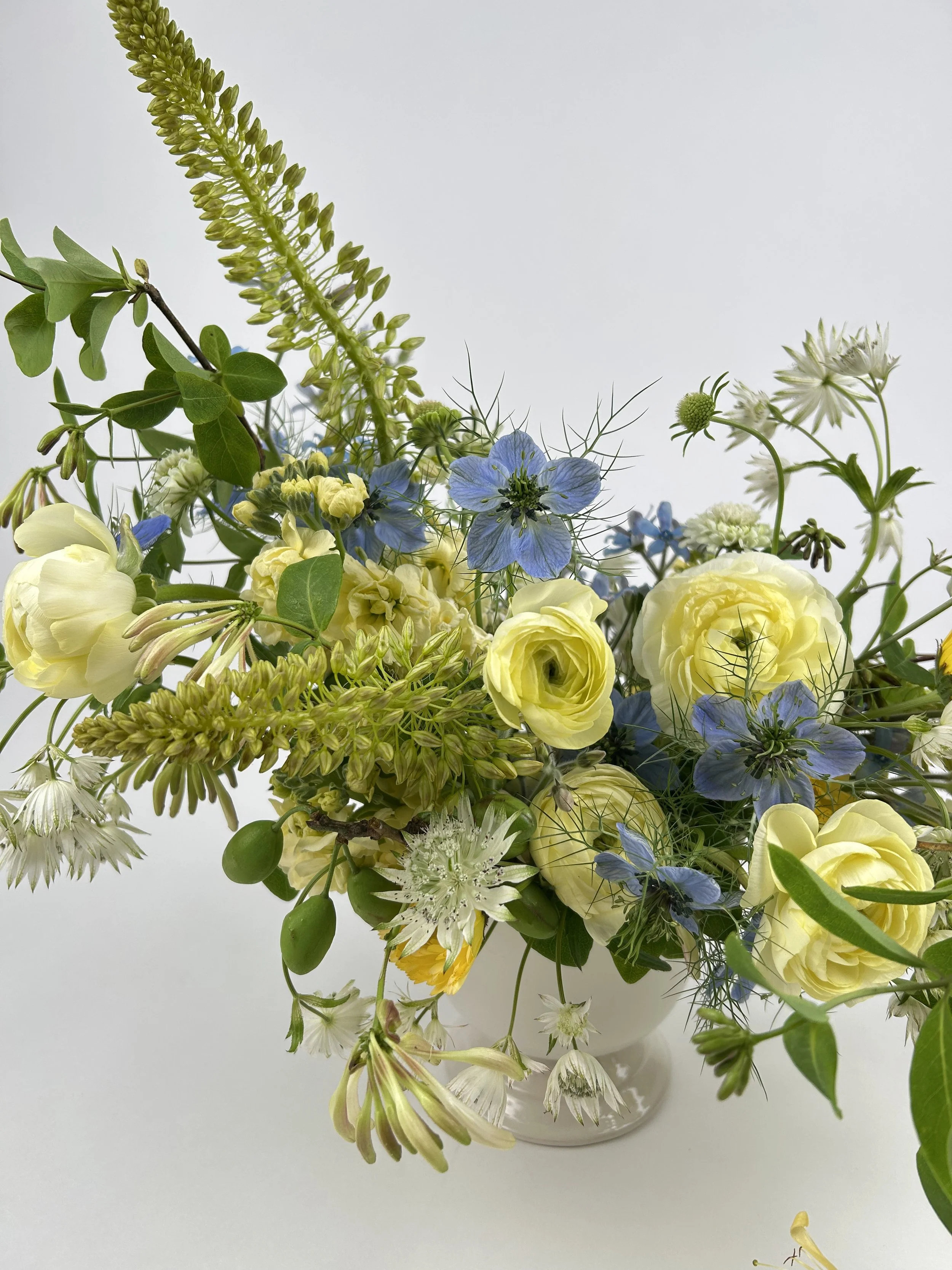 A floral arrangement with yellow and white flowers, interspersed with blue flowers and green foliage, in a white ceramic vase against a plain light background.