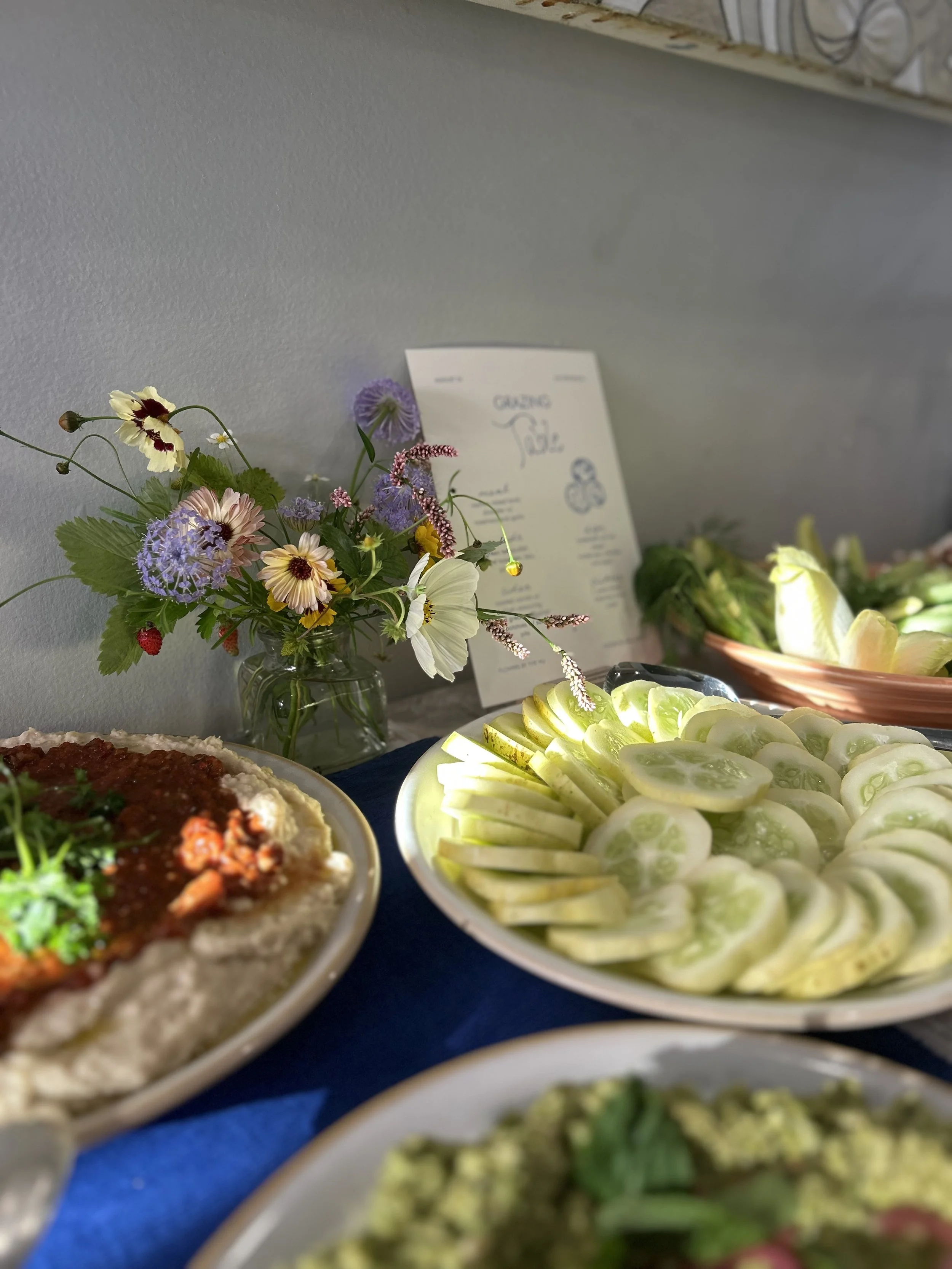 A buffet table with a platter of sliced cucumbers, a dish of chili with beans, and a bowl of salad garnished with herbs, alongside a vase of colorful wildflowers against a gray wall.