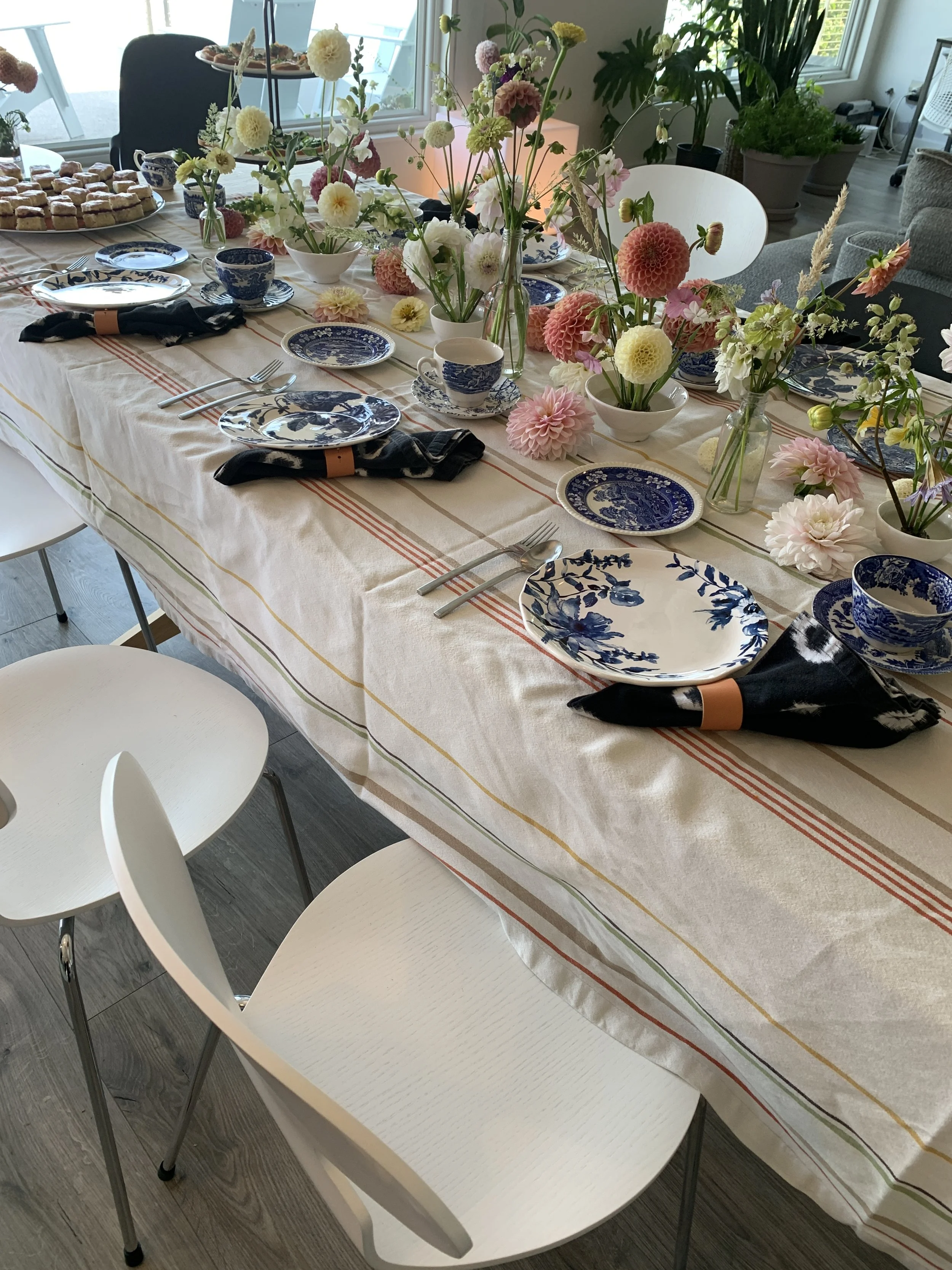A dining table set with floral-patterned blue and white dishes, black napkins with tan rings, silverware, and decorated with colorful fresh flowers in glass vases. The table is covered with a white tablecloth with red, yellow, and green lines, and su
