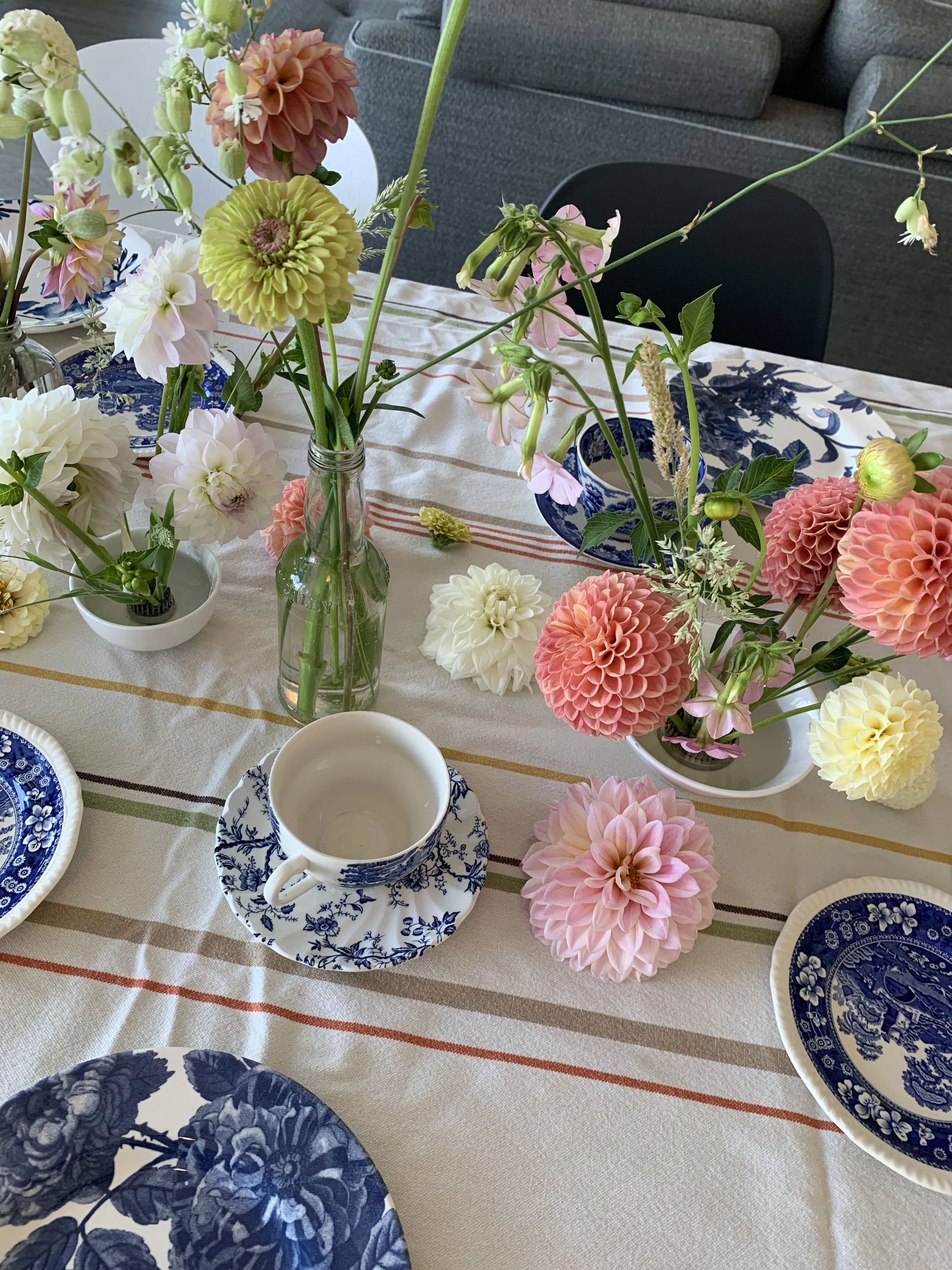 A table decorated with colorful flowers in vases, along with blue and white patterned plates and a white teacup. The tablecloth has pastel stripes, and the background includes a gray sofa and chairs.