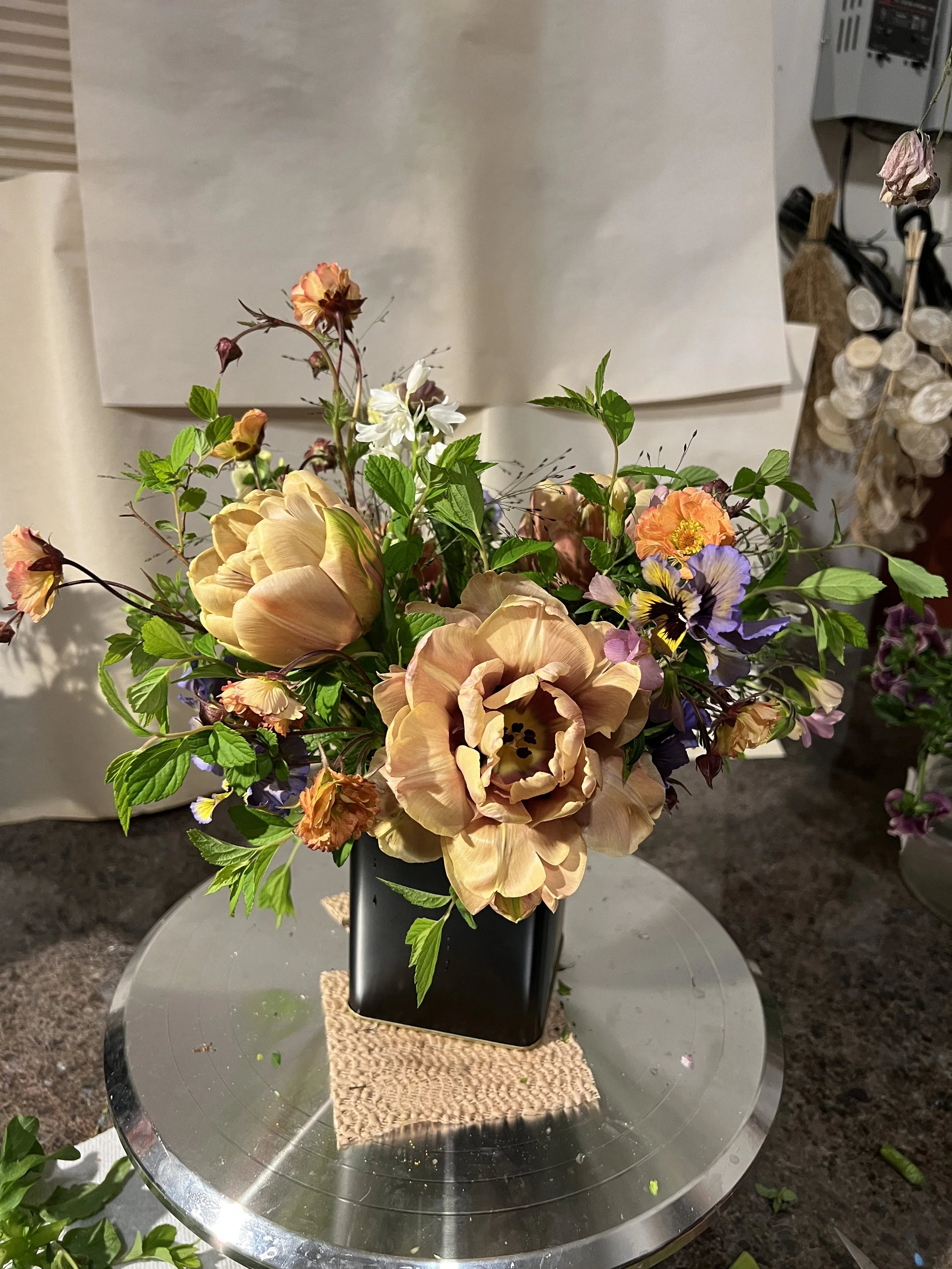 A flower arrangement in a black vase on a round, reflective metal table with a textured beige mat underneath. The arrangement includes large beige peonies, smaller peach and purple flowers, and green leaves. The background features a plain white wall