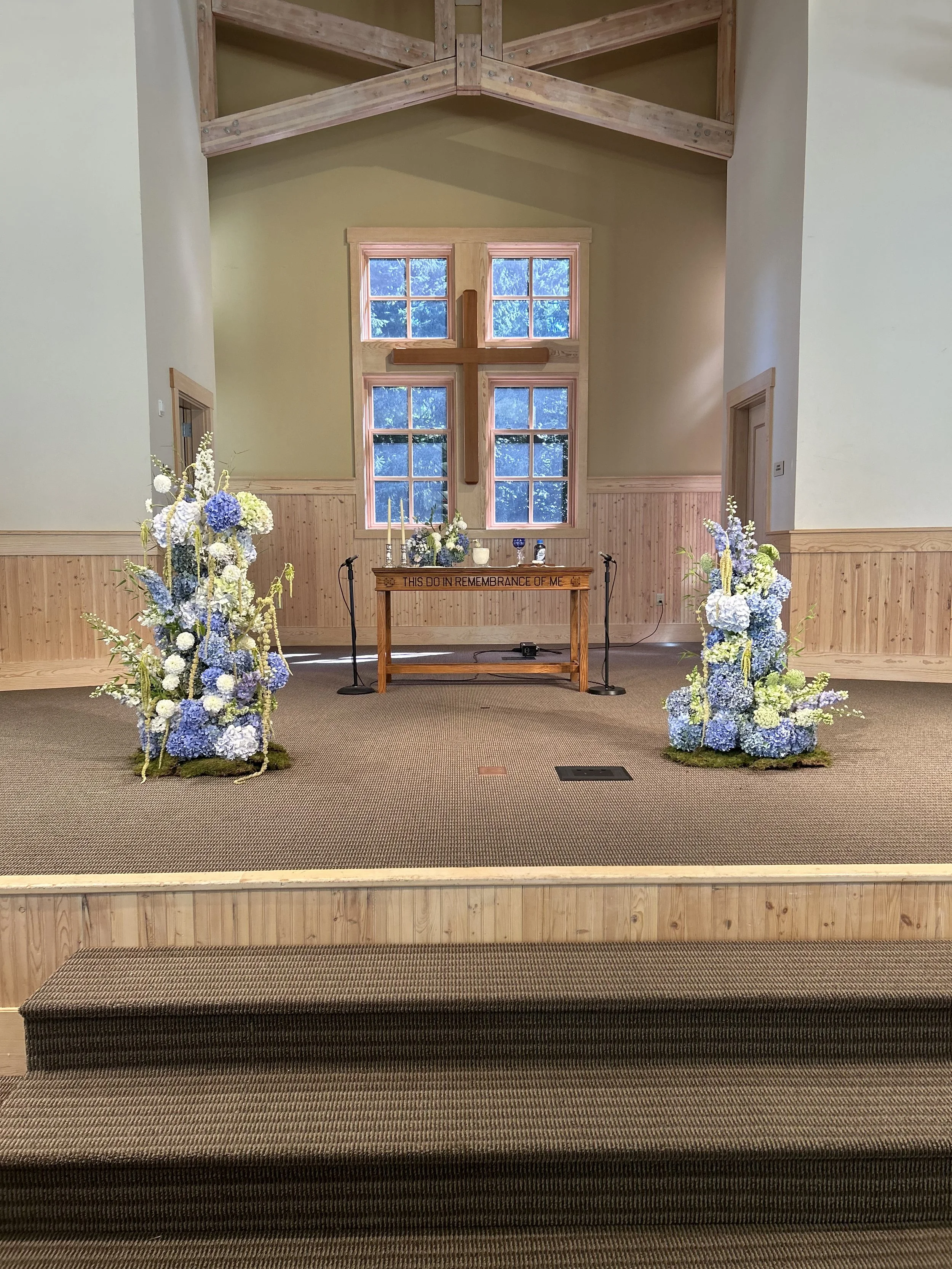 Interior of a chapel with floral arrangements, a wooden altar, a large wooden cross on the back wall, and windows clustered around it.