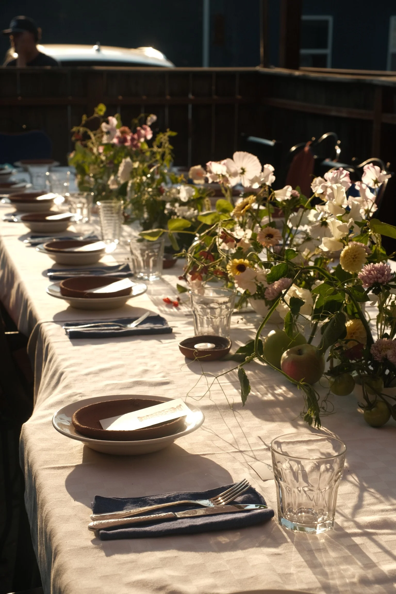 A long table set for a meal with white tablecloth, plates, utensils, and glasses, decorated with a large floral centerpiece in natural sunlight.