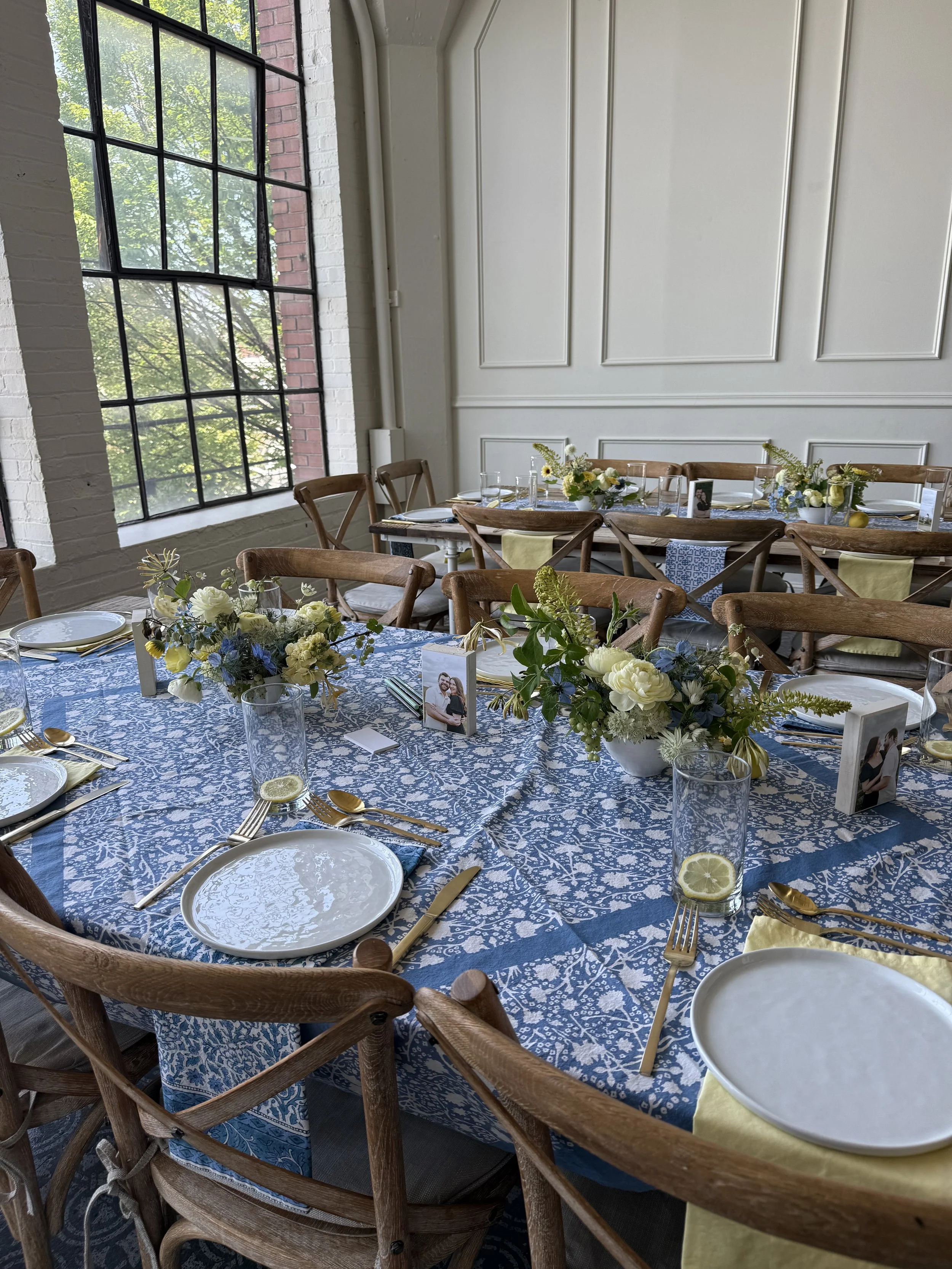 A decorated event table with floral centerpieces, white and blue tableware, and framed photos in a bright room with large windows and wooden chairs.
