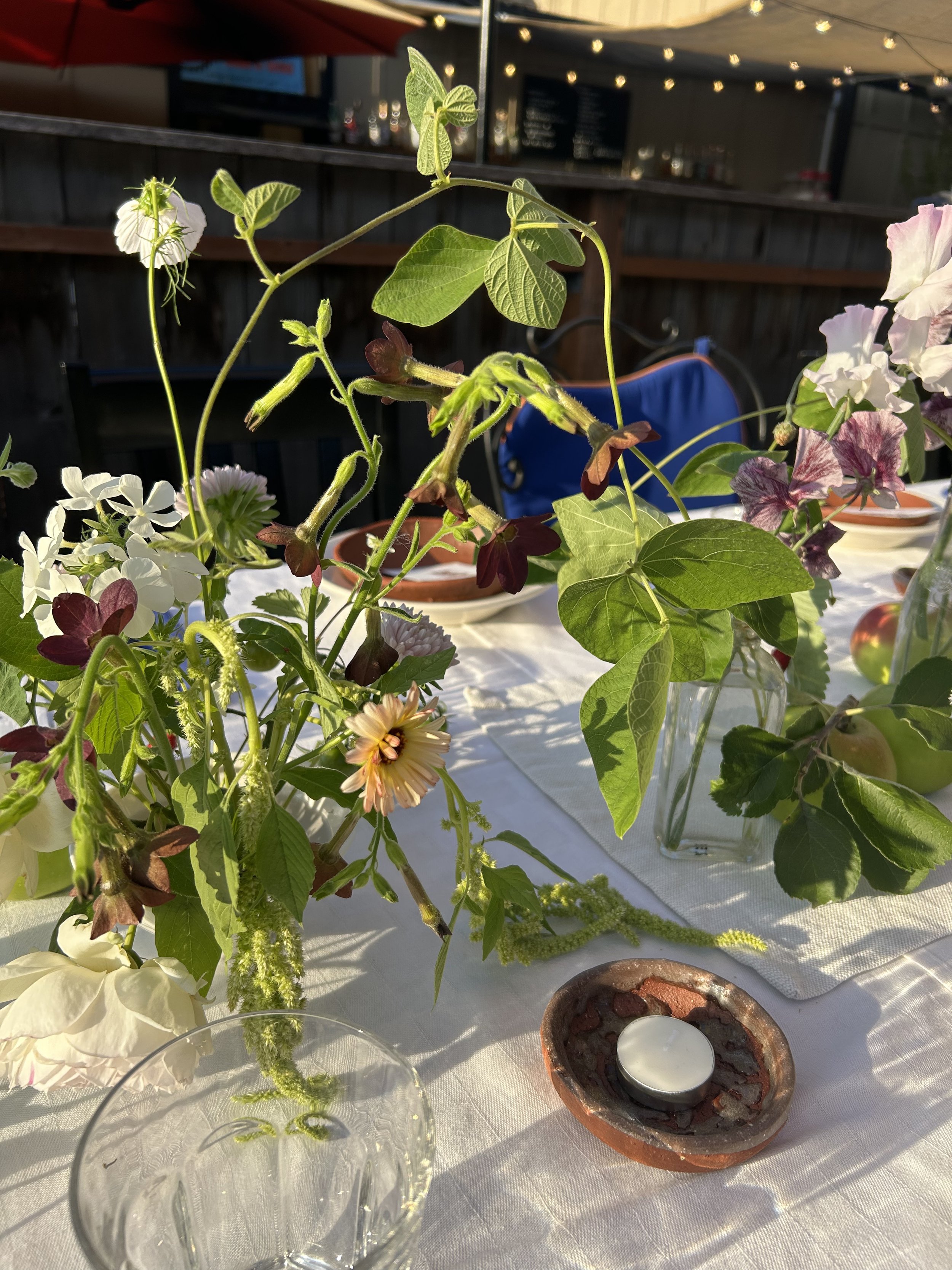 Arrangement of potted flowers and greenery on a table with a candle holder, in an outdoor setting with string lights overhead.