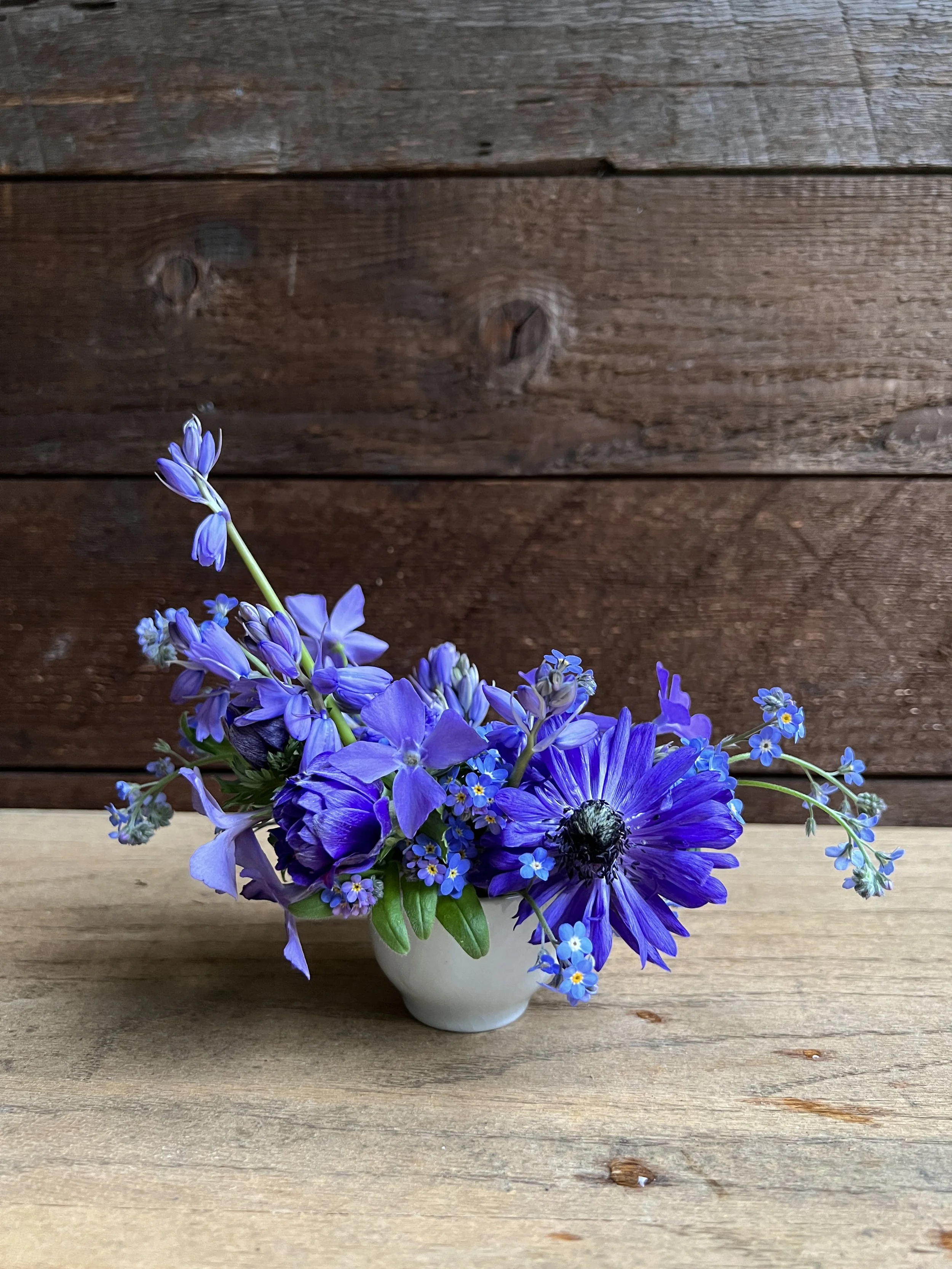 Small white cup with a bouquet of purple and blue flowers, including daisies and lavender, on a wooden surface against a wooden background.