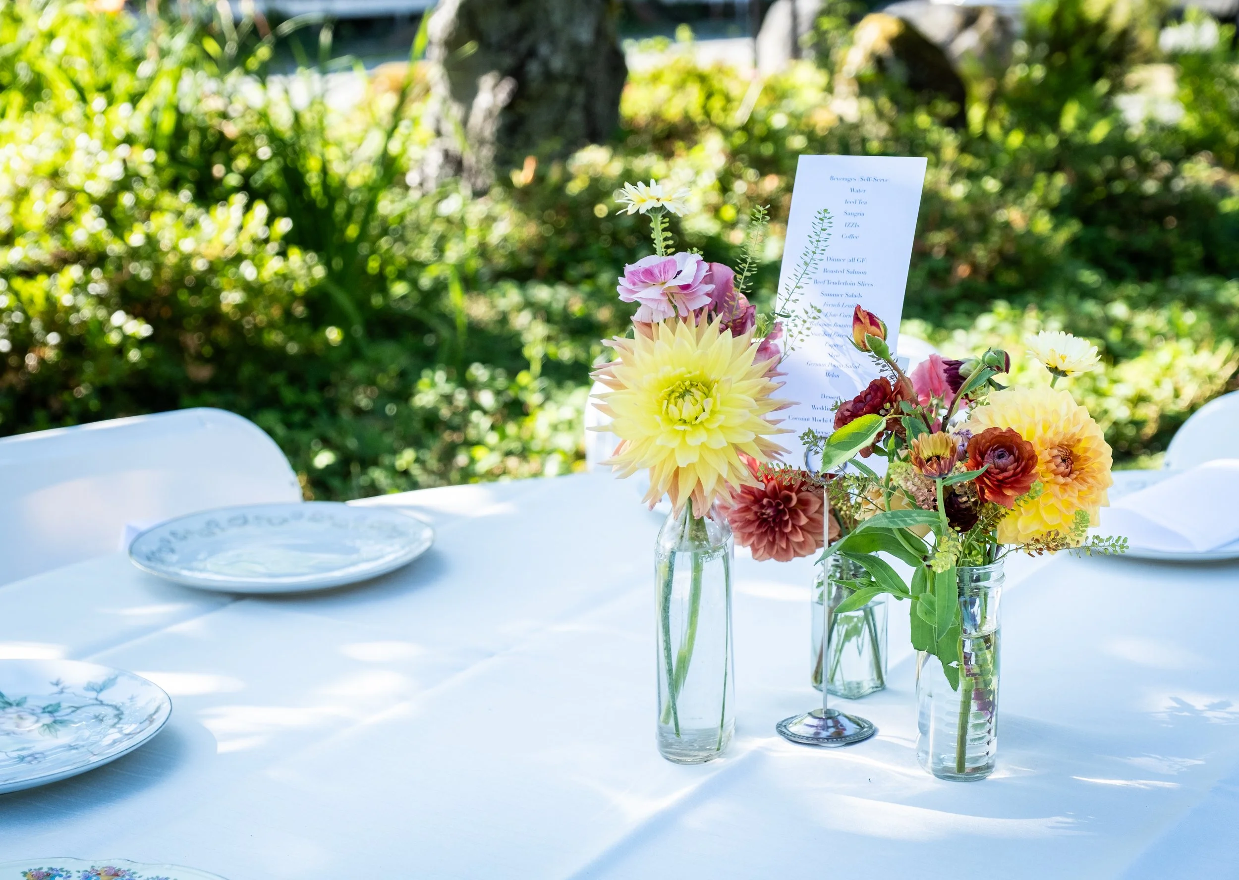 Table set outdoors with white tablecloth, floral centerpieces in glass vases, and white plates, with greenery in the background.