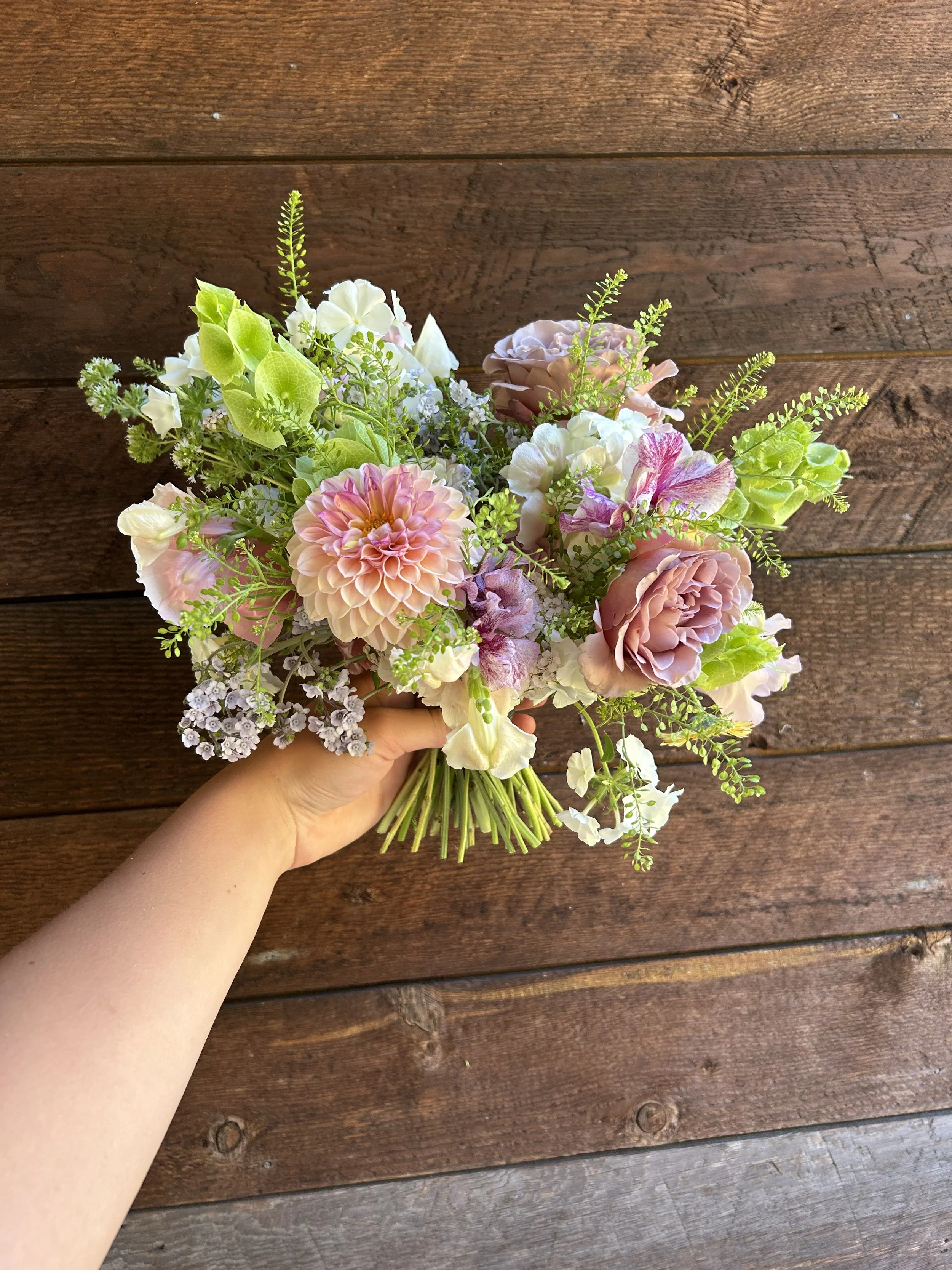 Hand holding a colorful bridal bouquet of pink, purple, and white flowers against a wooden floor background.