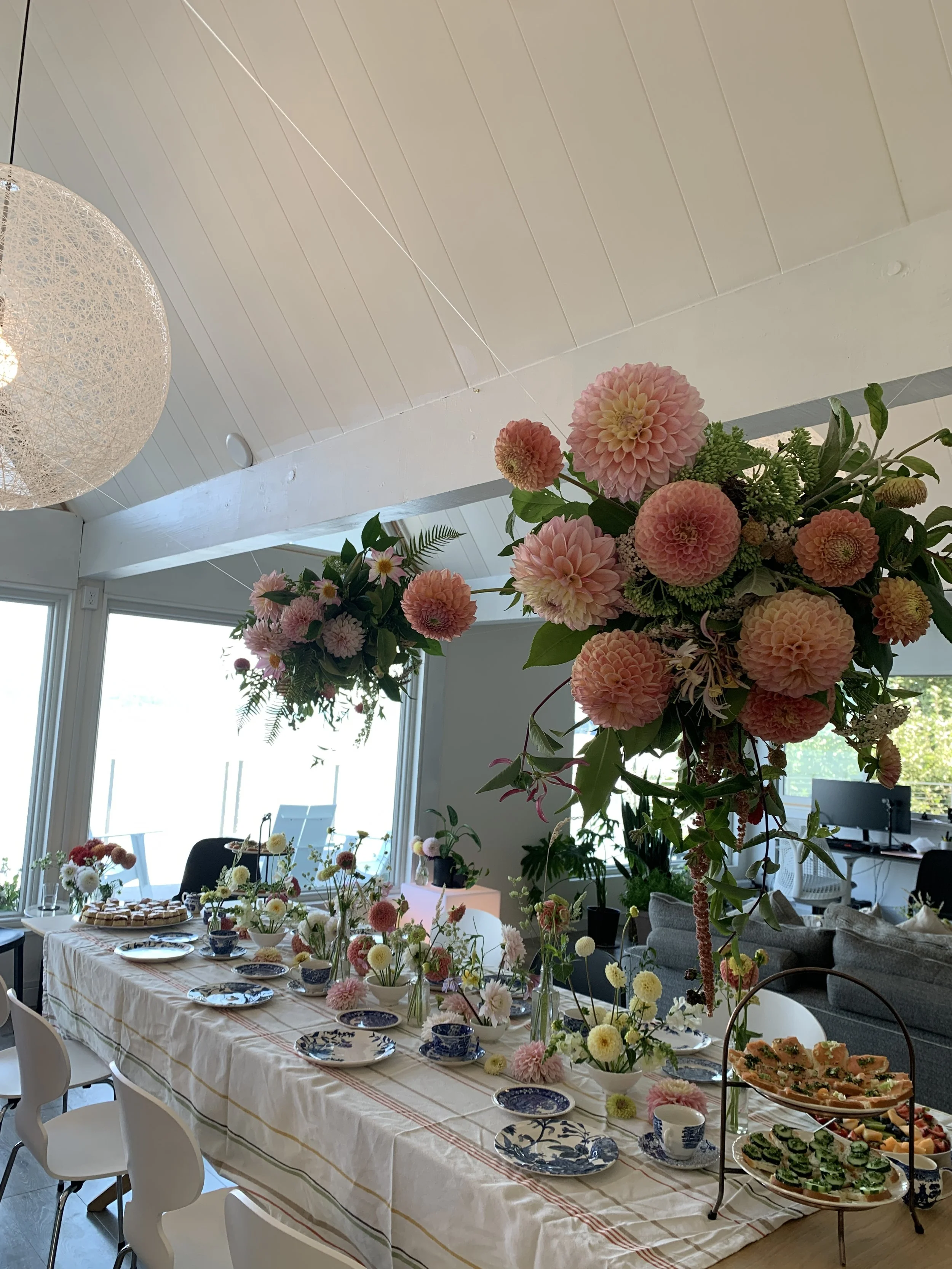 A decorated dining table set for a celebration in a bright room with large windows, floral arrangements with pink and white flowers, and a tiered tray with snacks or appetizers.