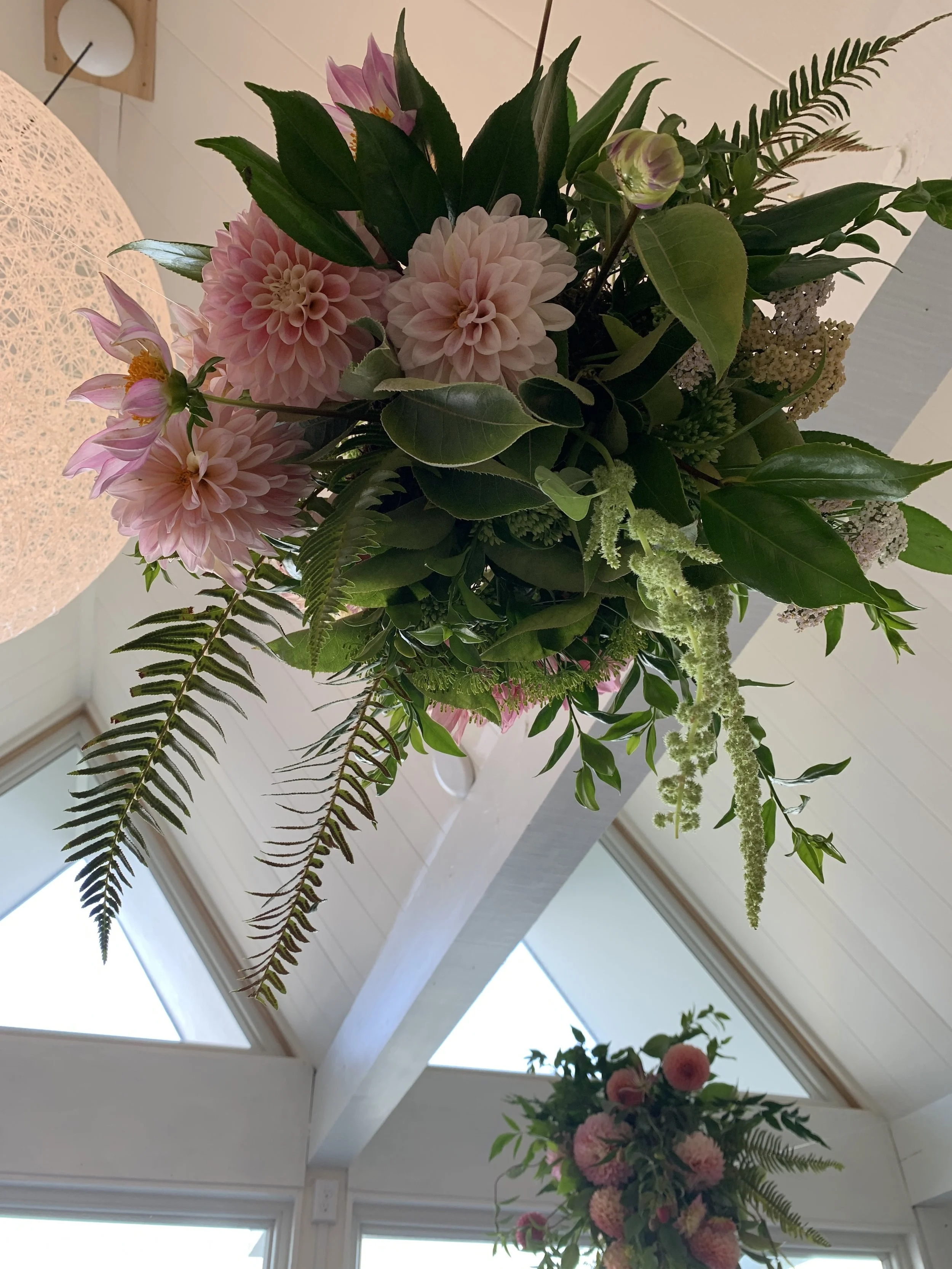 Decorative floral arrangement with pink dahlias, lilies, ferns, and green leaves in a vase, placed on a table near a ceiling with skylights.