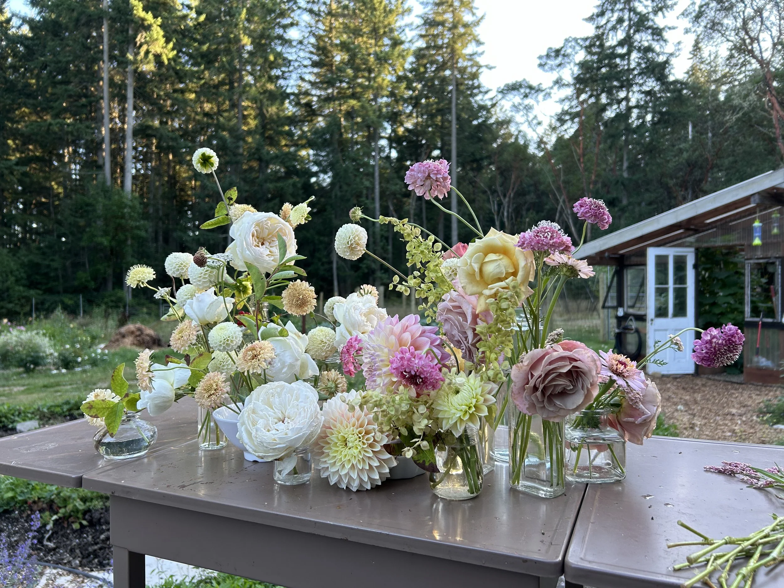 Floral arrangement of white, pink, lavender, and cream-colored flowers on a wooden table outdoors with trees and a barn in the background.